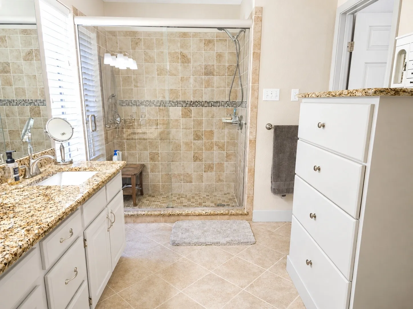 Tiled shower enclosure with glass surround, granular countertop on white cabinetry, and tiled flooring