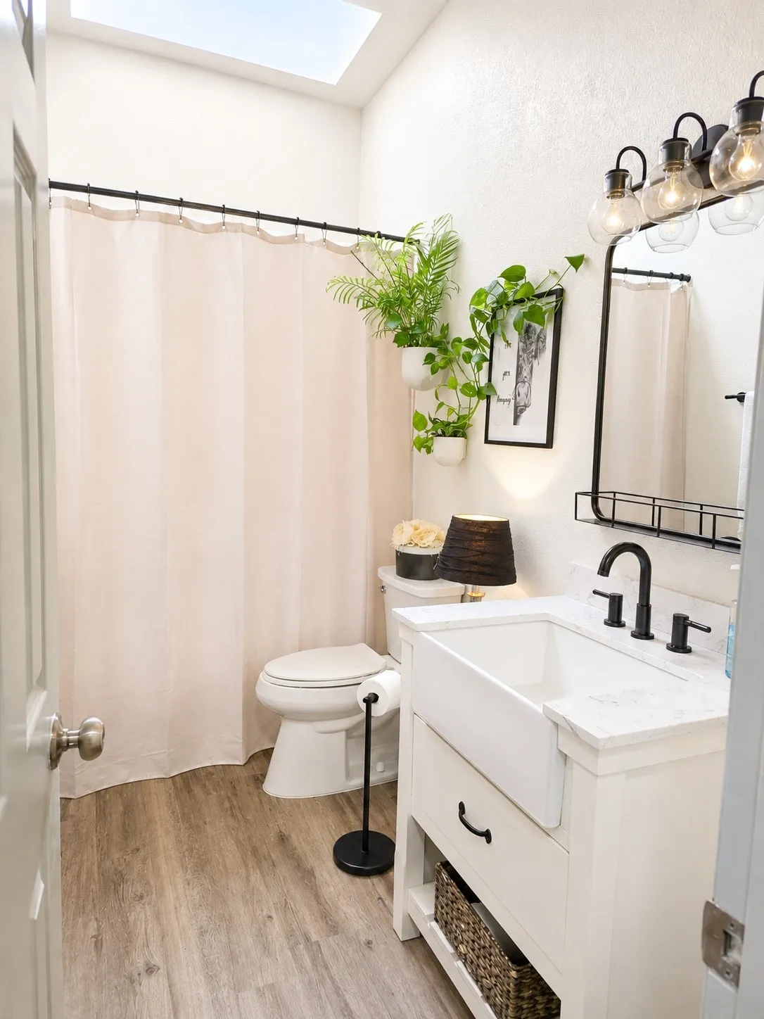 Bathroom featuring a skylight, wood-finish flooring, a white porcelain farmhouse sink, a white vanity with black hardware, and a black-framed mirror with industrial-style lighting
