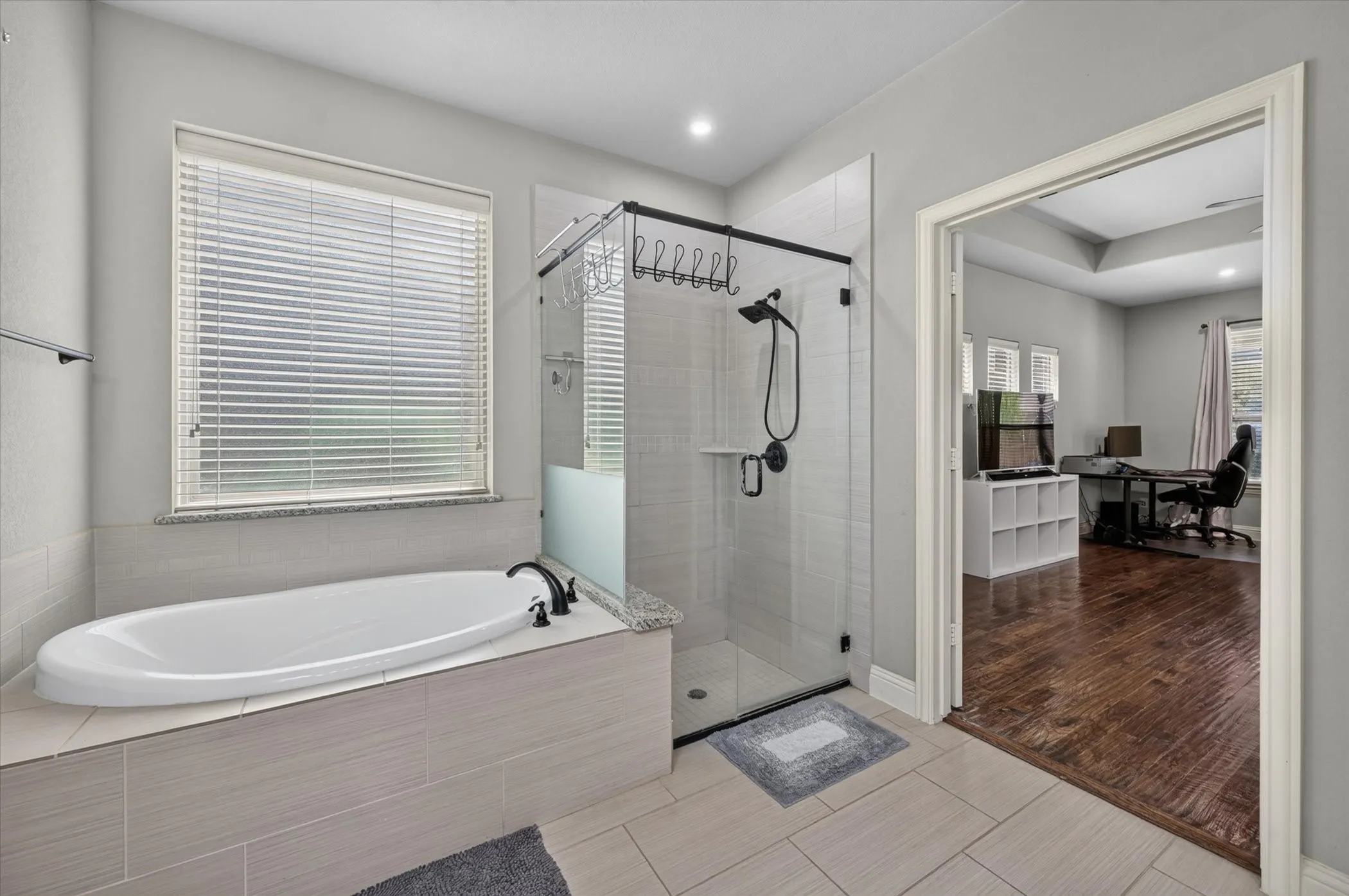 Soaking tub with black fixtures, tile-surround shower with glass enclosure, white window blinds, and recessed lighting