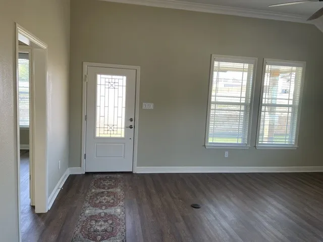 Vaulted ceiling with crown molding, decorative glass front door, two windows with blinds, wood-finish flooring, and a ceiling fan