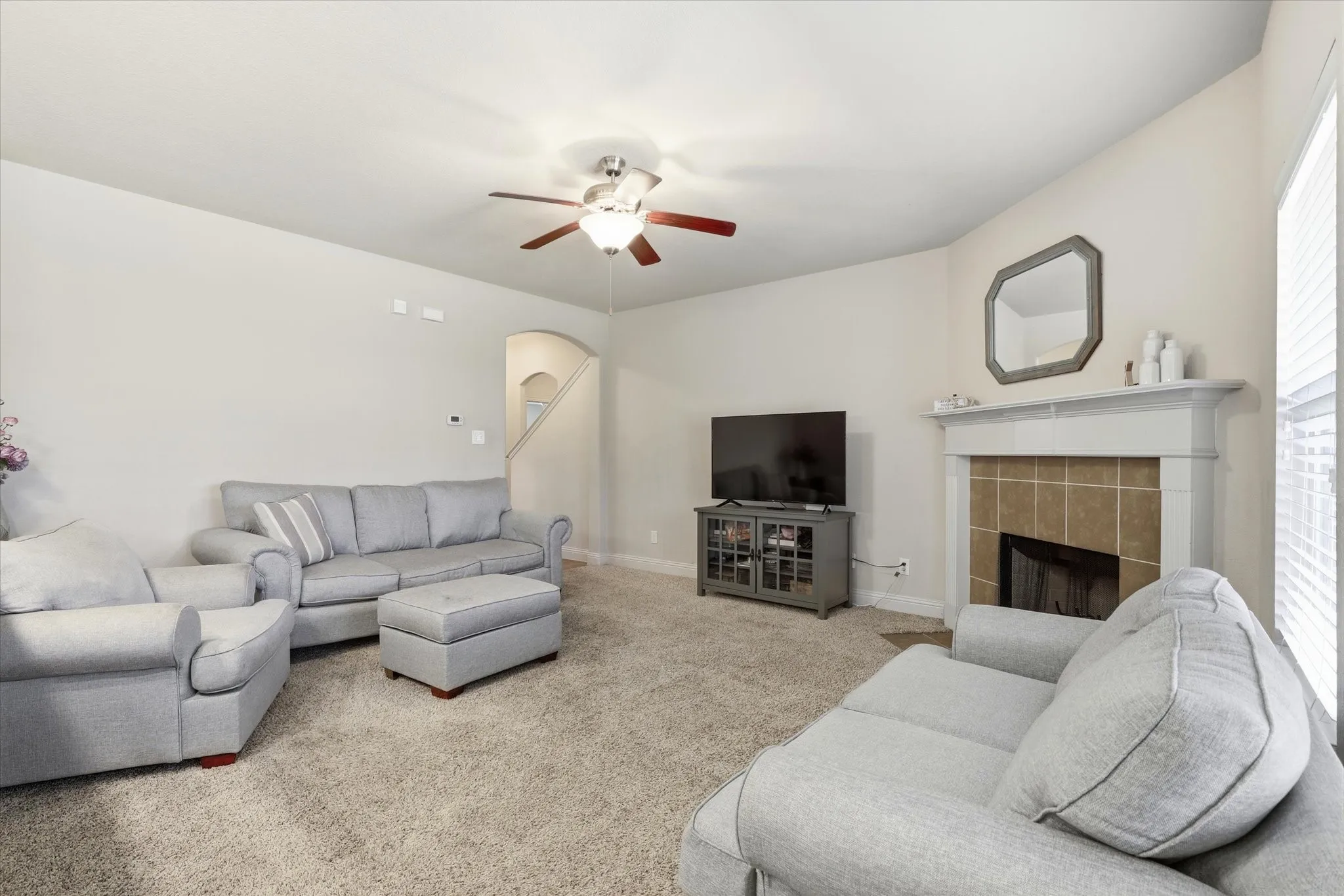 Carpeted living area featuring a ceiling fan with integrated lighting, a fireplace with a tile surround and substantial mantel, and an arched doorway leading to a staircase