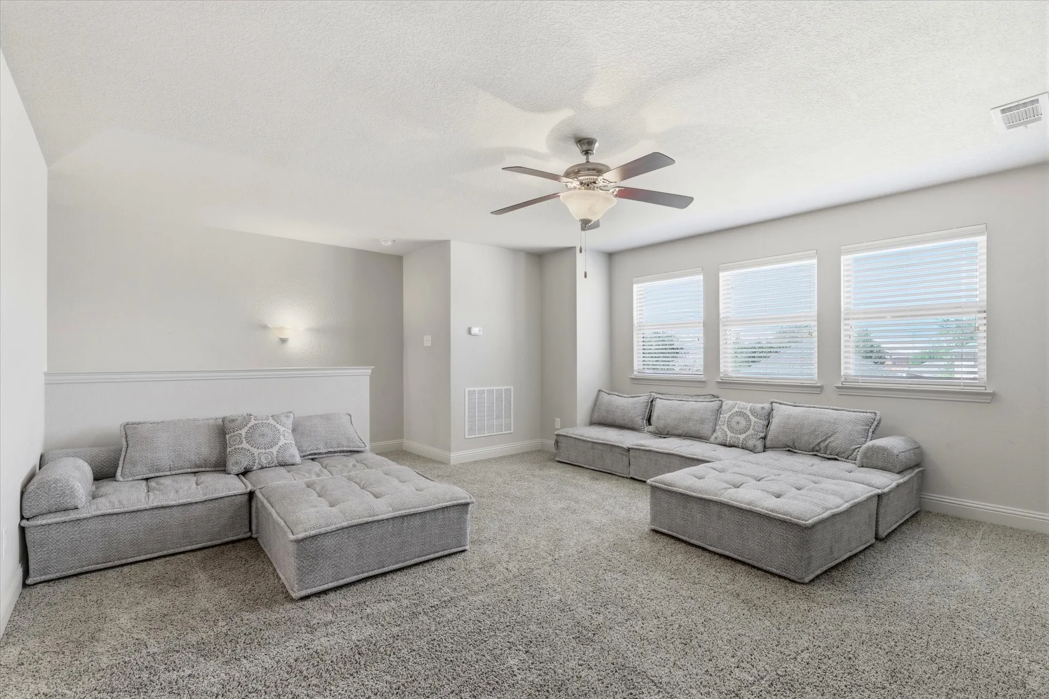 Spacious room featuring carpeting, multiple windows with blinds, a ceiling fan with integrated lighting, and wall-mounted sconces