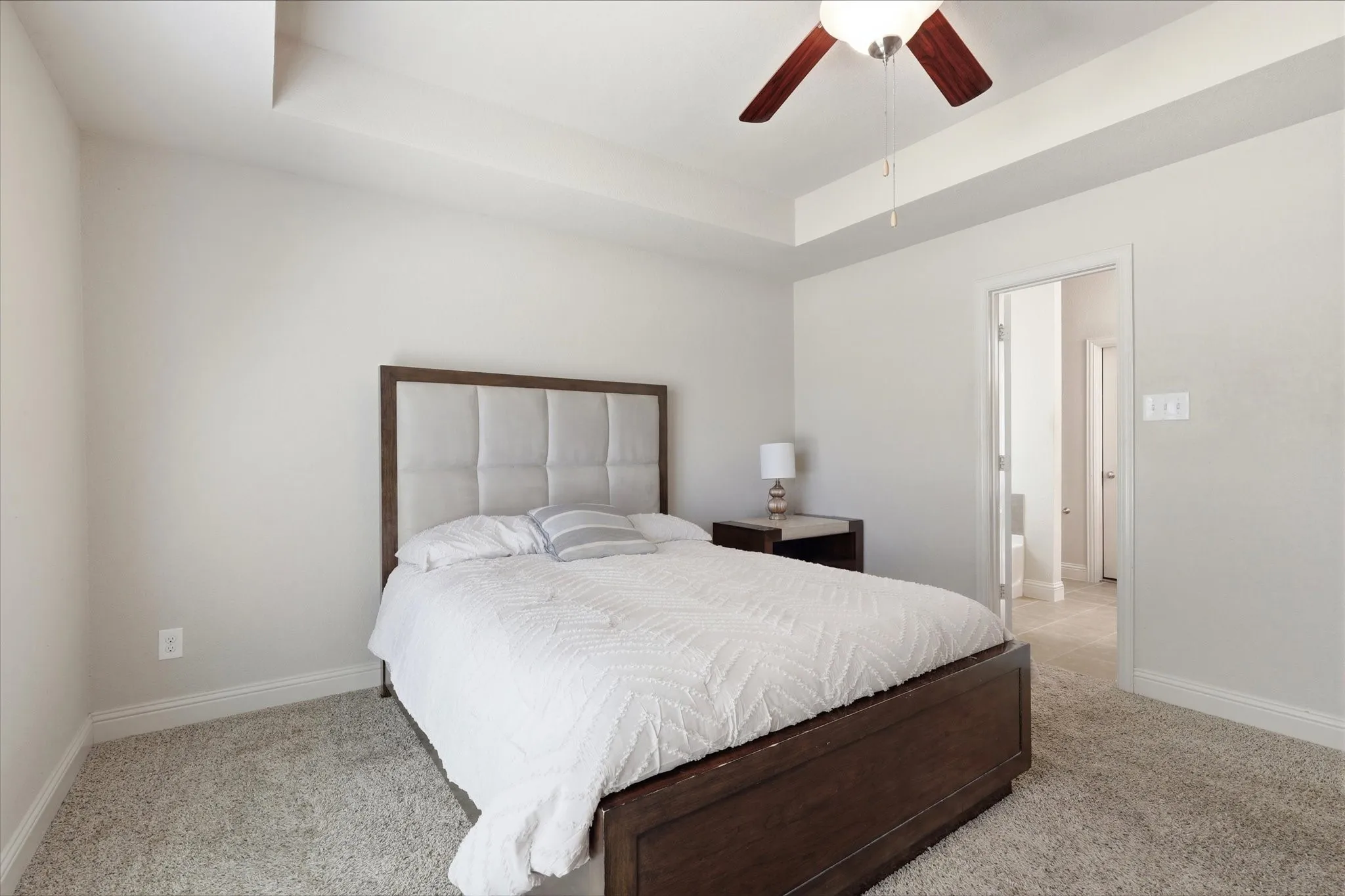 Tray ceiling with a ceiling fan, light gray carpeting, and a doorway leading to an additional space