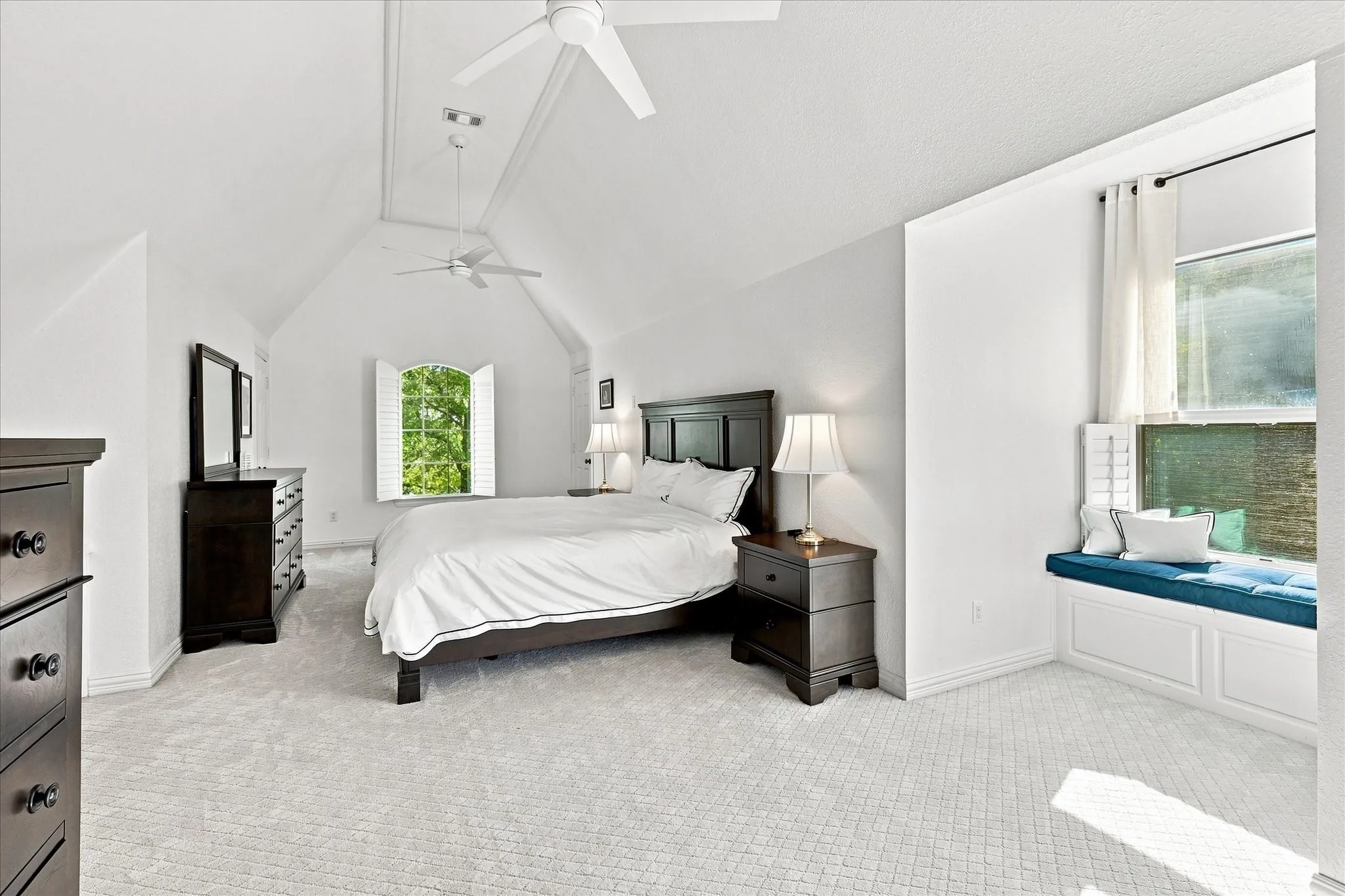 Vaulted ceiling with dual ceiling fans, window with plantation shutters, built-in window seat with storage, and neutral carpet flooring