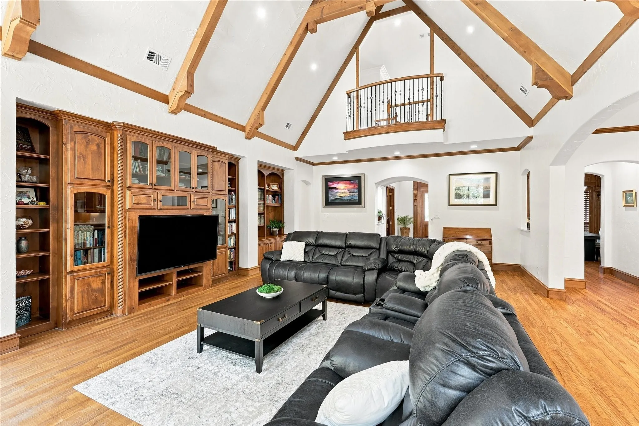 Vaulted white ceilings with exposed wood beams, wood-finish flooring, built-in wooden media cabinetry, arched doorways, and an interior balcony with wrought iron railing