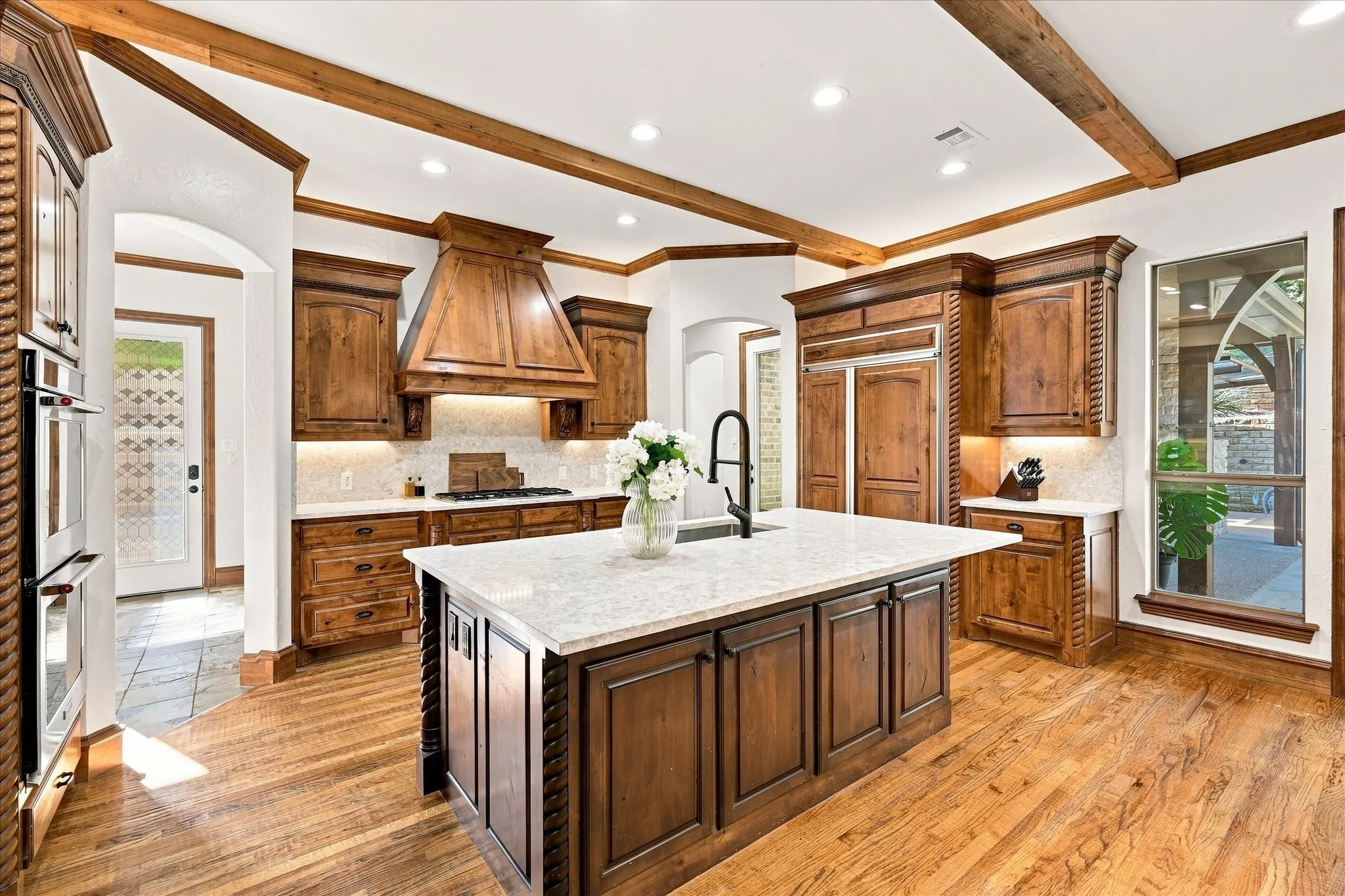 Gourmet kitchen featuring wood-finish flooring, a large center island with a stone countertop, wood cabinetry, stainless steel double ovens, and decorative wood ceiling beams