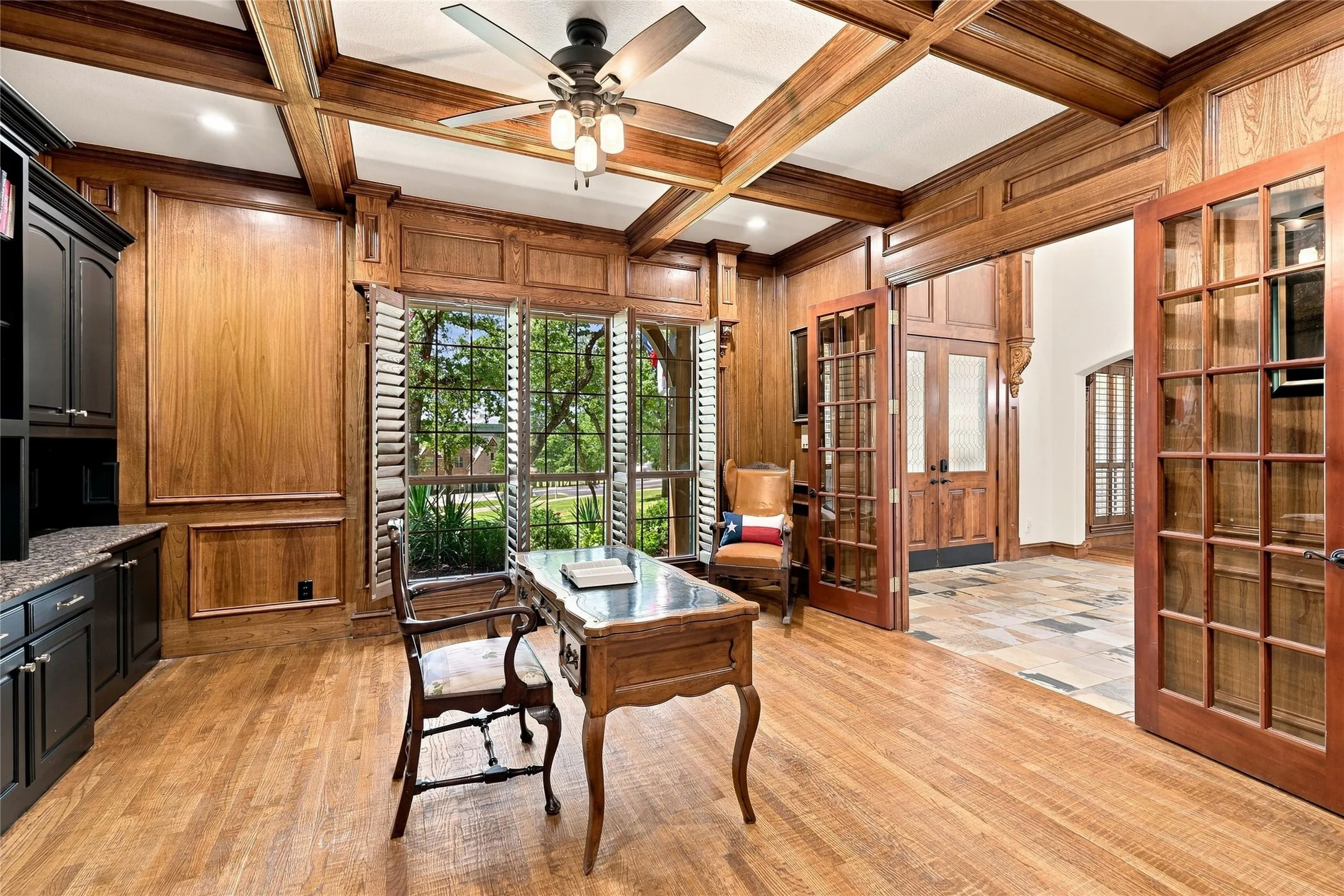 Hardwood flooring, wood-paneled walls, coffered ceiling, wood-framed French doors, and built-in cabinetry with stone countertops