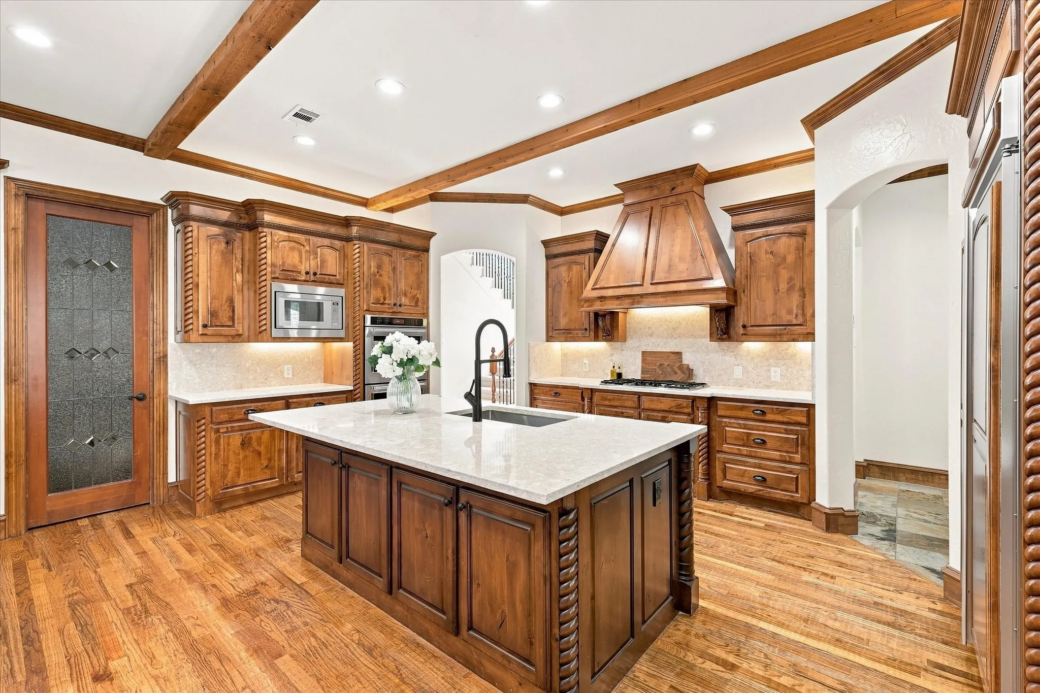 Gourmet kitchen featuring wood-finish flooring, a central island with a light-toned countertop, ample custom wood cabinetry, stainless steel appliances, and exposed ceiling beams