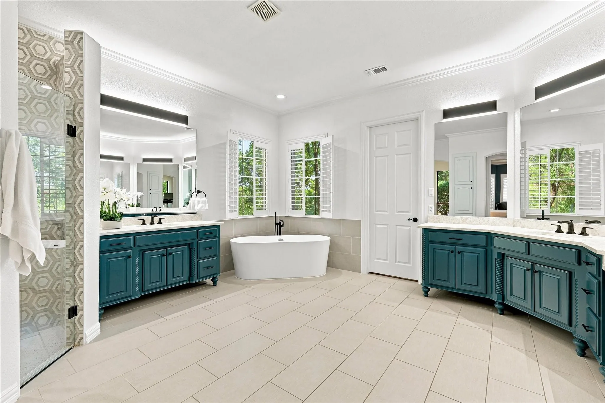 Freestanding soaking tub, two individual vanities with integrated sinks, glass-enclosed shower with hexagonal tile, shuttered windows, and light tile flooring