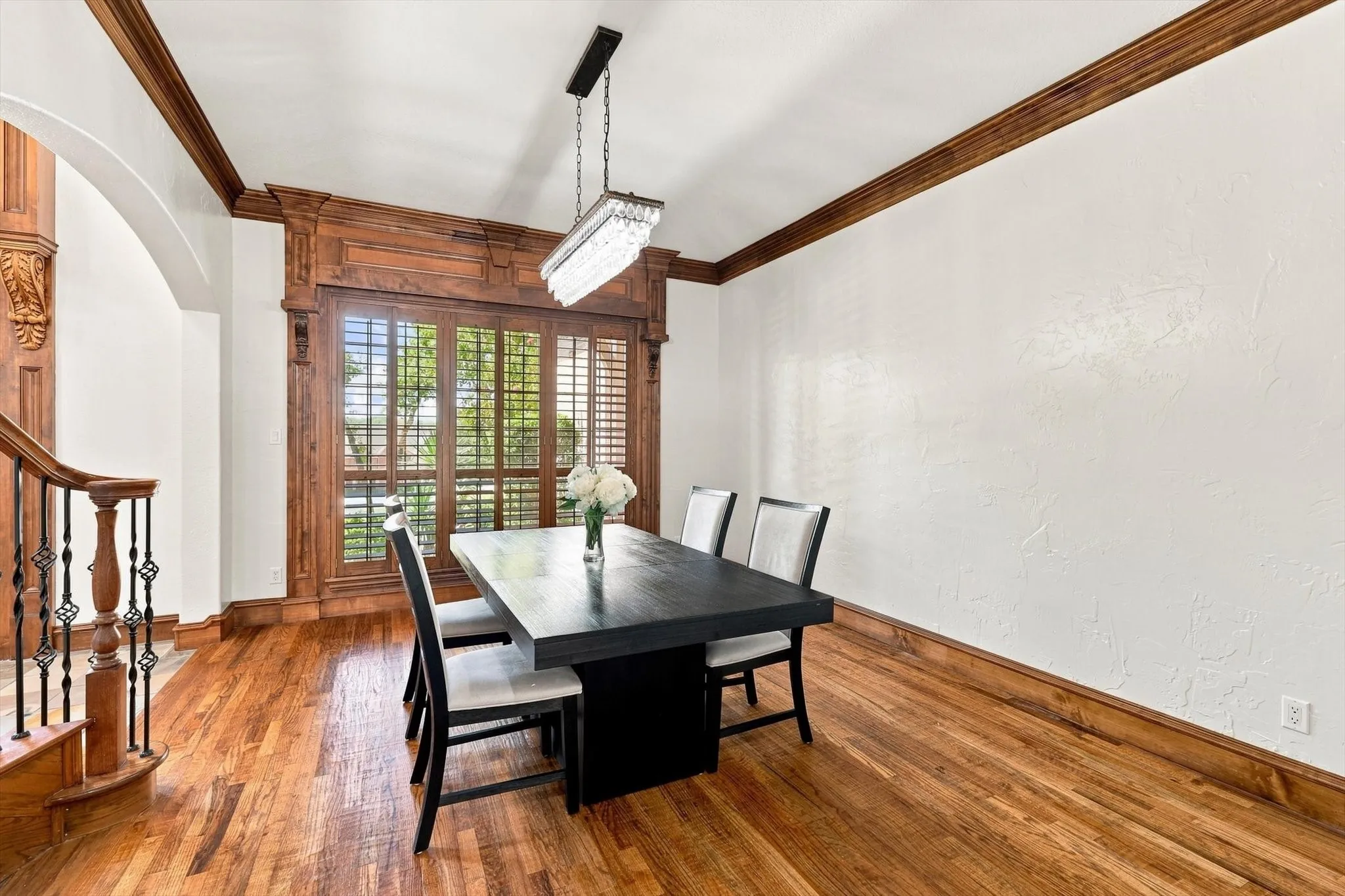 Hardwood flooring, ornate wood-finish window trim, plantation shutters, wrought iron stair railing, and a modern rectangular chandelier