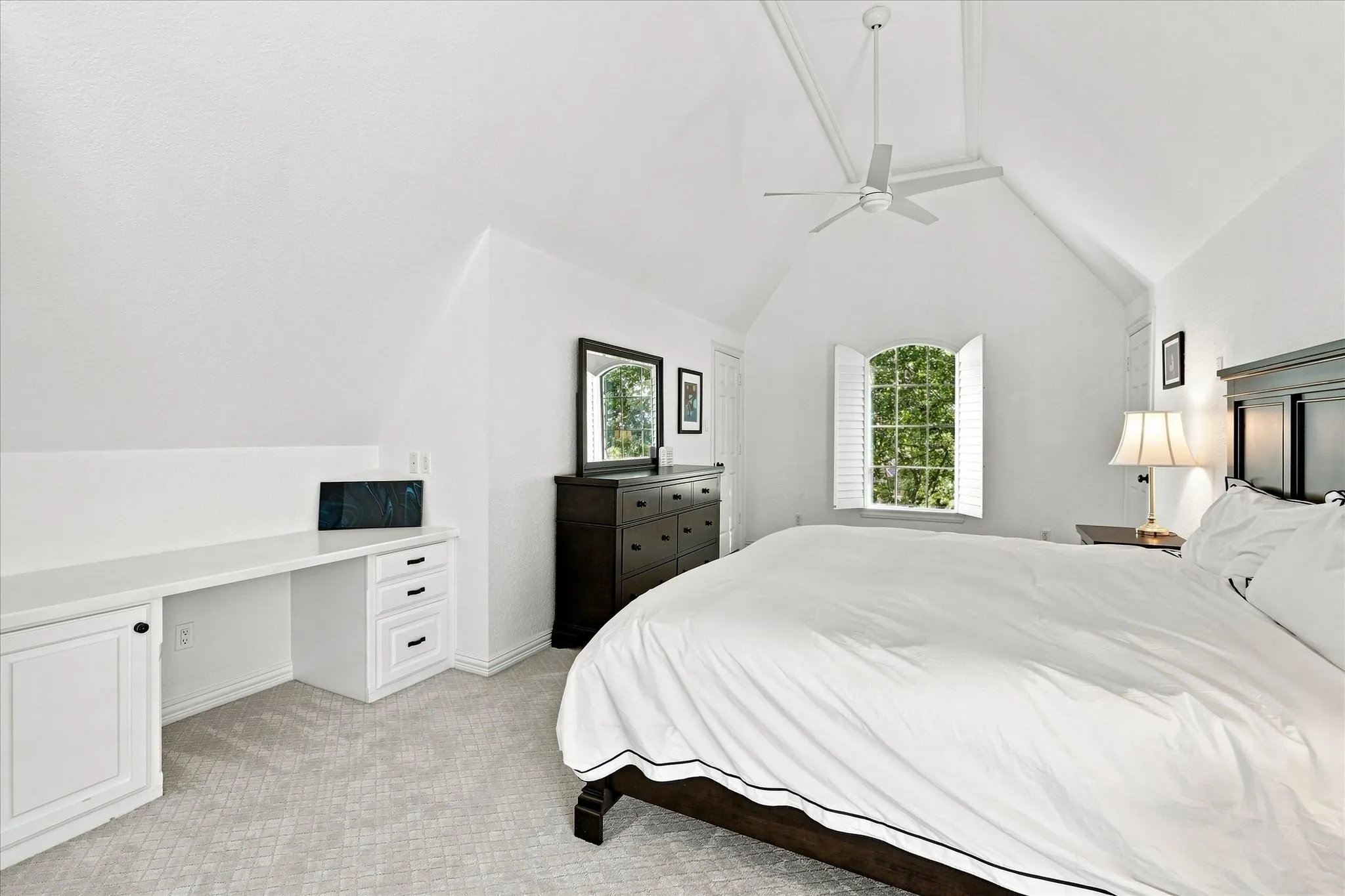 Vaulted ceiling, built-in desk, window with plantation shutters, carpet flooring, and a ceiling fan