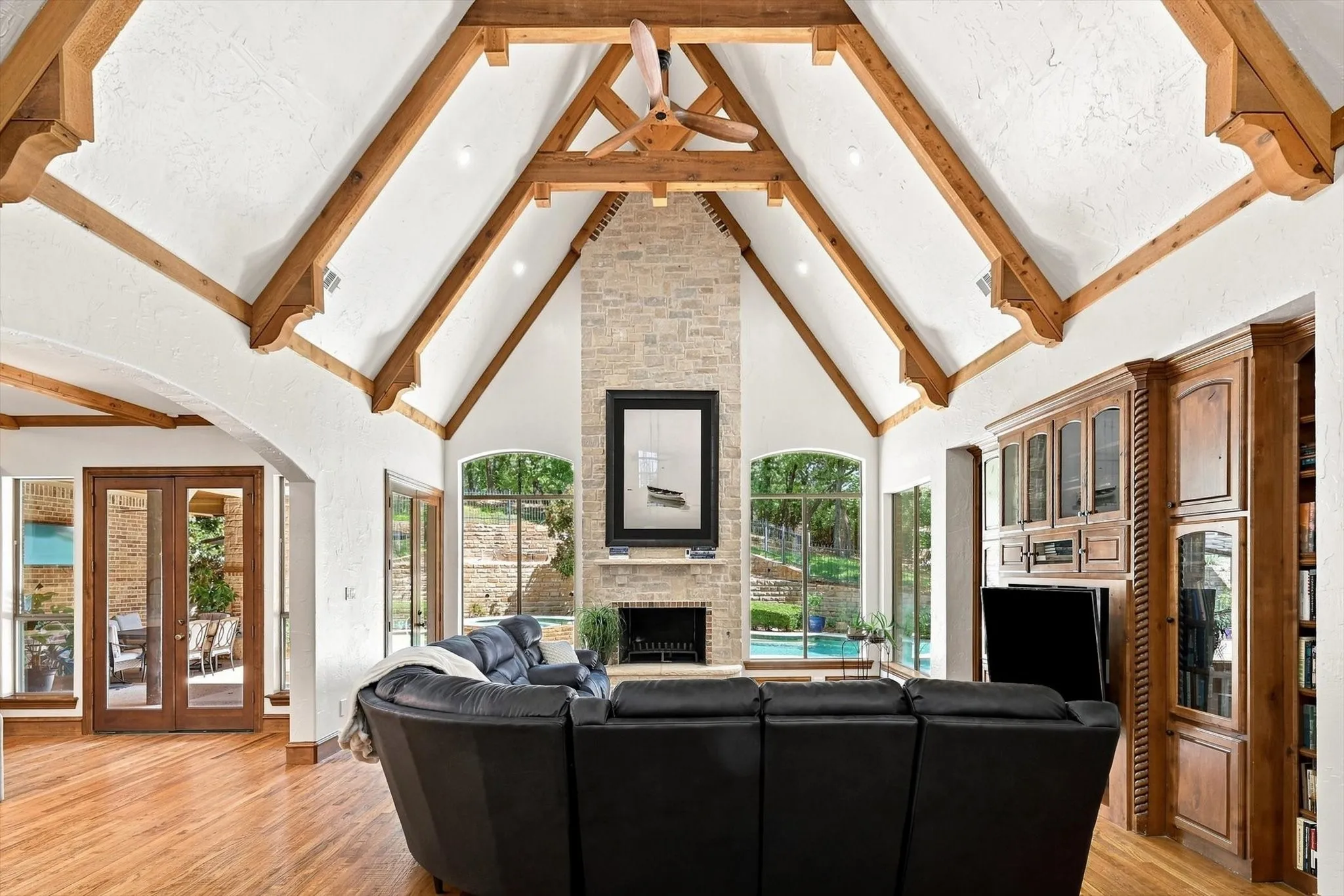 Vaulted ceiling with exposed wood beams, stone fireplace, built-in cabinetry, wood-finish flooring, and arched windows