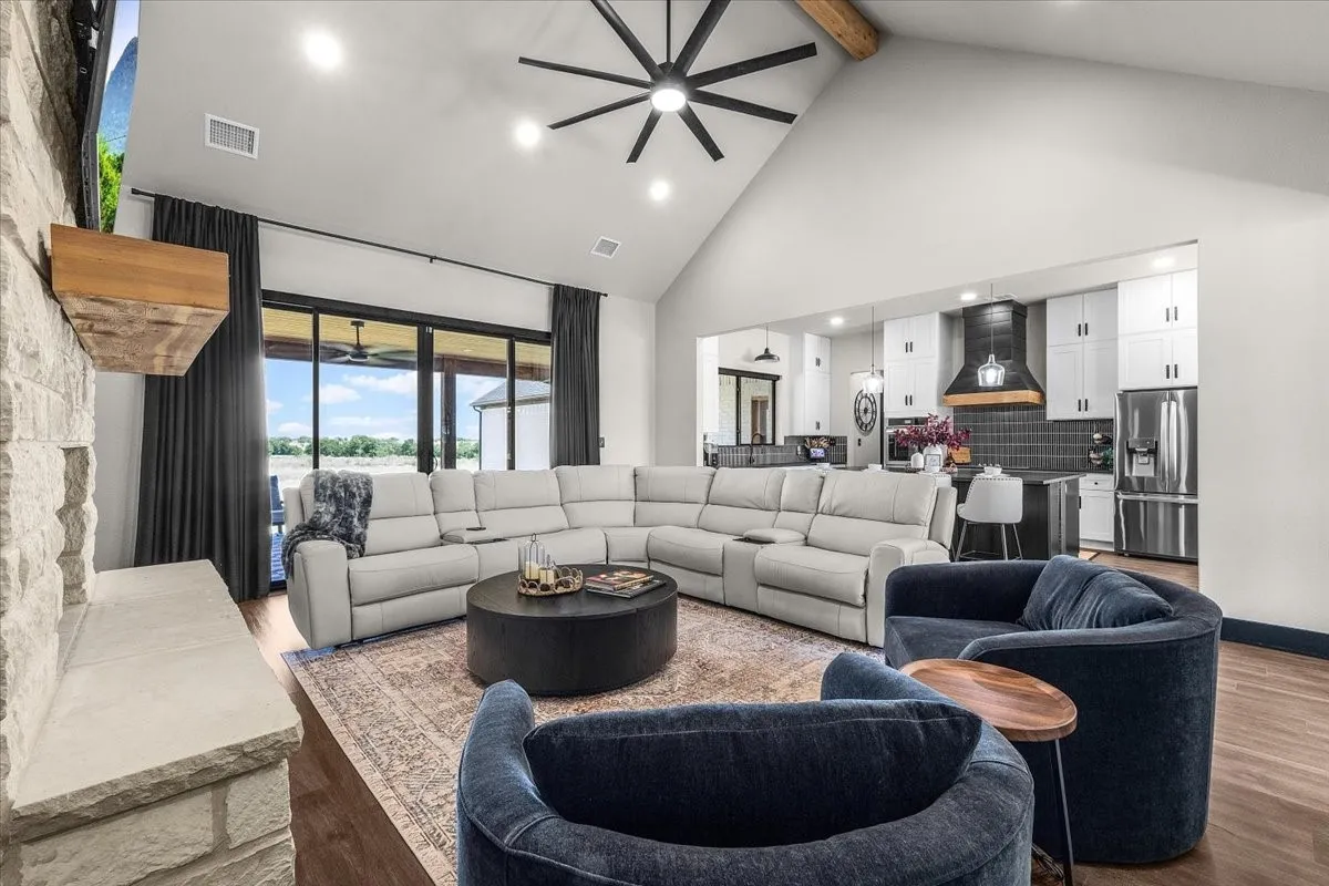 Vaulted ceiling with an exposed wood beam, stone fireplace with a wood mantel, wood-finish flooring, sliding glass doors, and a kitchen with white cabinetry and stainless steel appliances