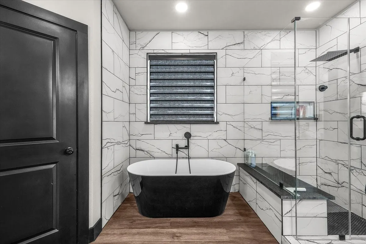 Freestanding soaking tub, wood-finish flooring, glass-enclosed shower, wall-mounted showerhead, and tiled walls with integrated shelving