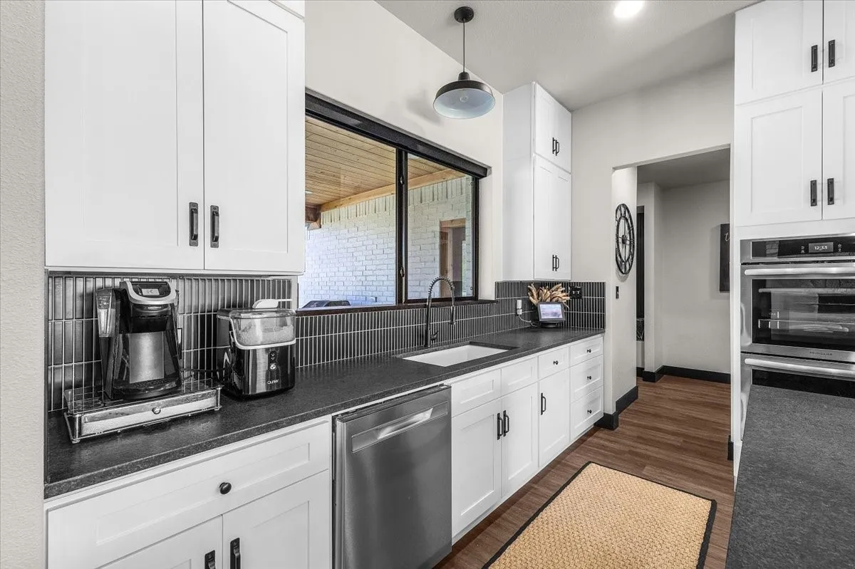 White cabinetry with contrasting dark hardware, stainless steel dishwasher, built-in double oven, dark tile backsplash, and dark countertops