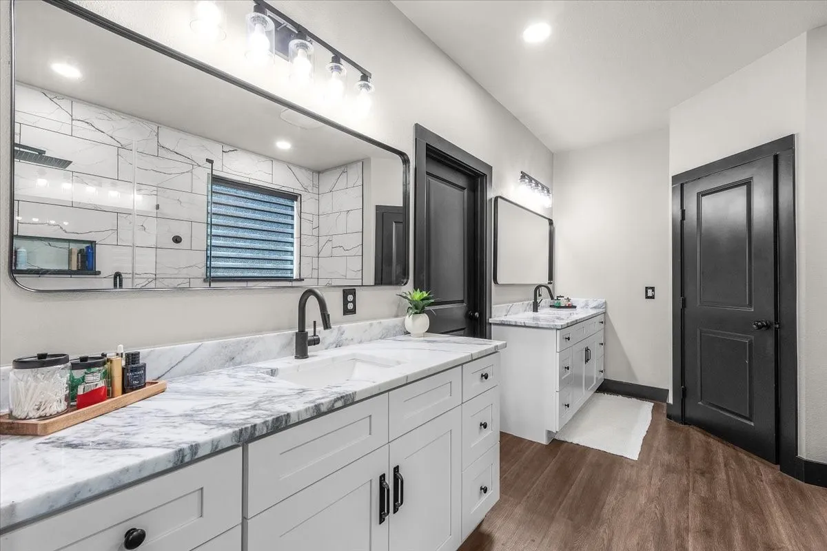 Dual white vanities with marble countertops, wood-finish flooring, black framed mirrors, and matte black fixtures