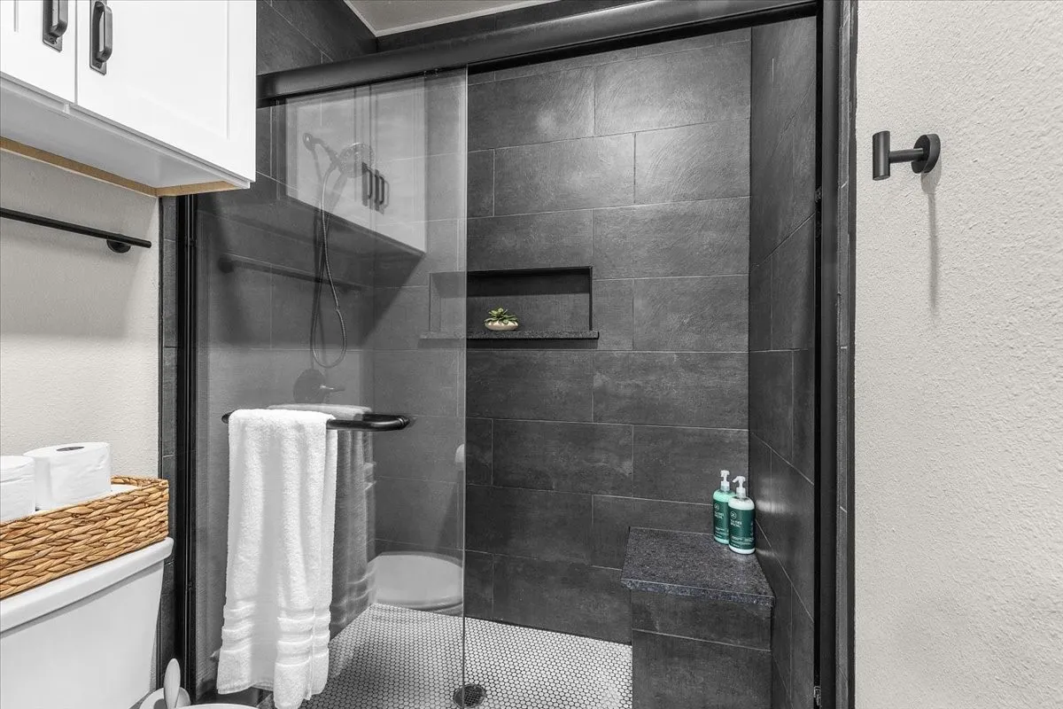 Glass enclosed shower featuring dark gray tile, built-in recessed shelf, built-in bench, and a rainfall shower head