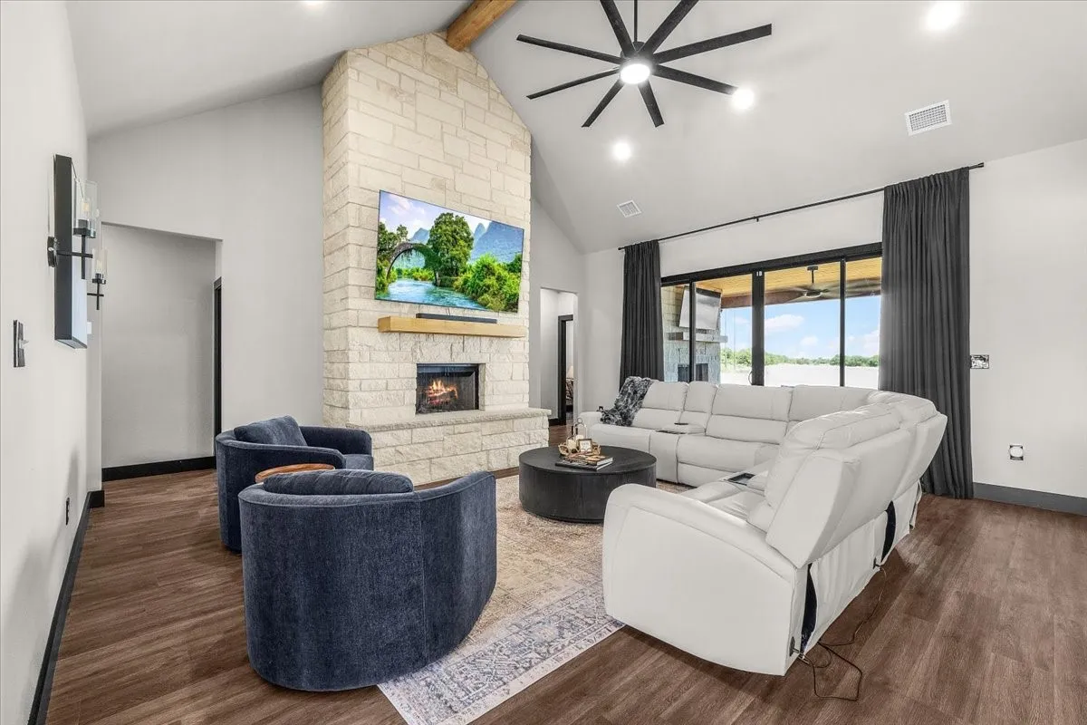 Vaulted ceiling with exposed wood beam, stone-clad fireplace, wood-finish flooring, dark-framed sliding glass doors, and recessed lighting