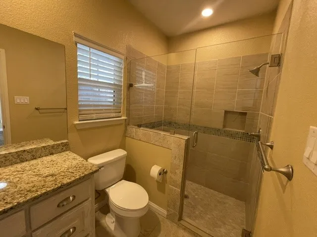 Granite-finish countertop with white cabinetry, glass-enclosed shower with tiled walls and an integrated niche, and a window with blinds