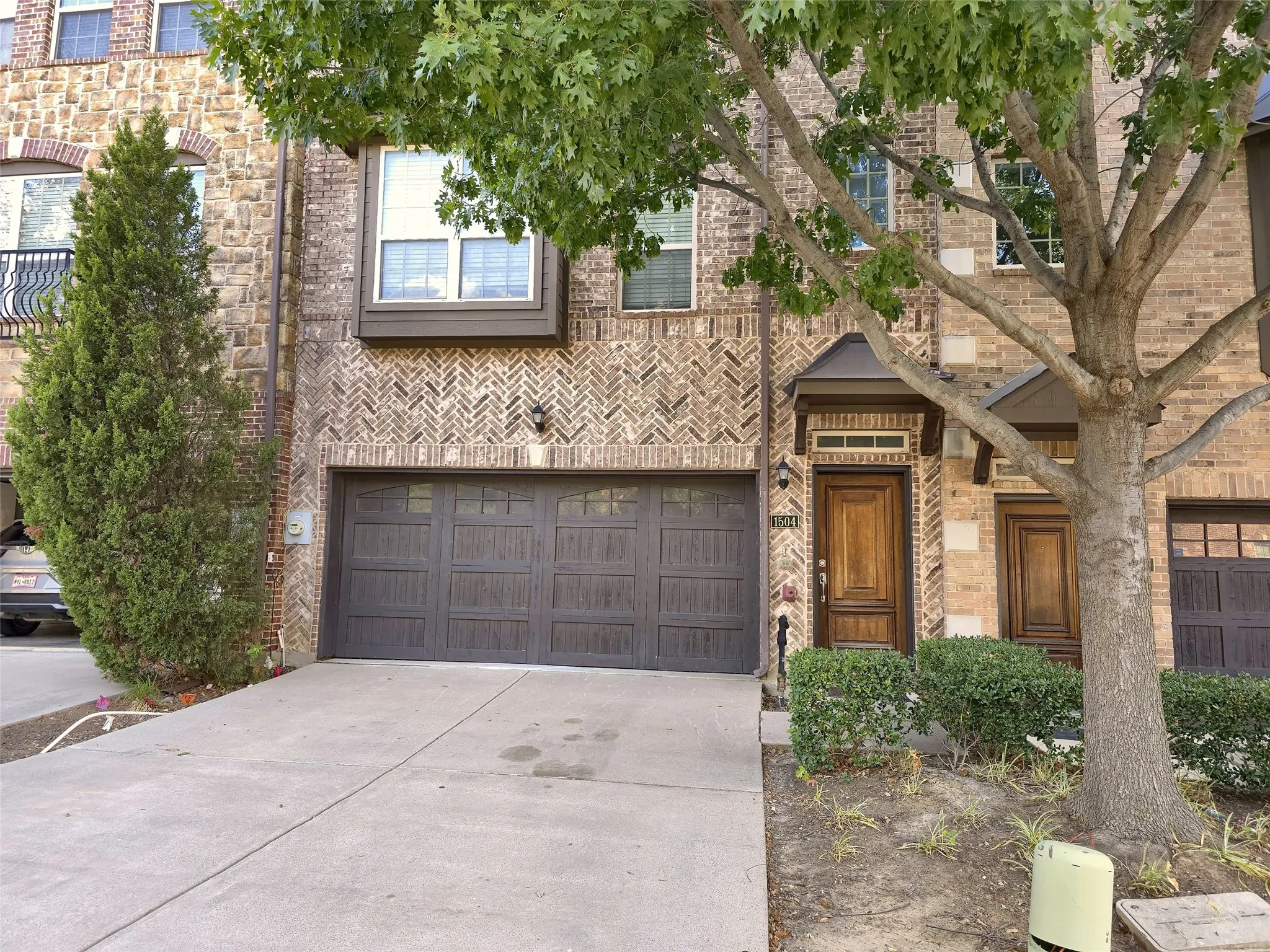 Brick facade featuring decorative herringbone patterns, wood-paneled garage door, arched brick window accents, natural wood entry door, and a second-story balcony with wrought iron railing