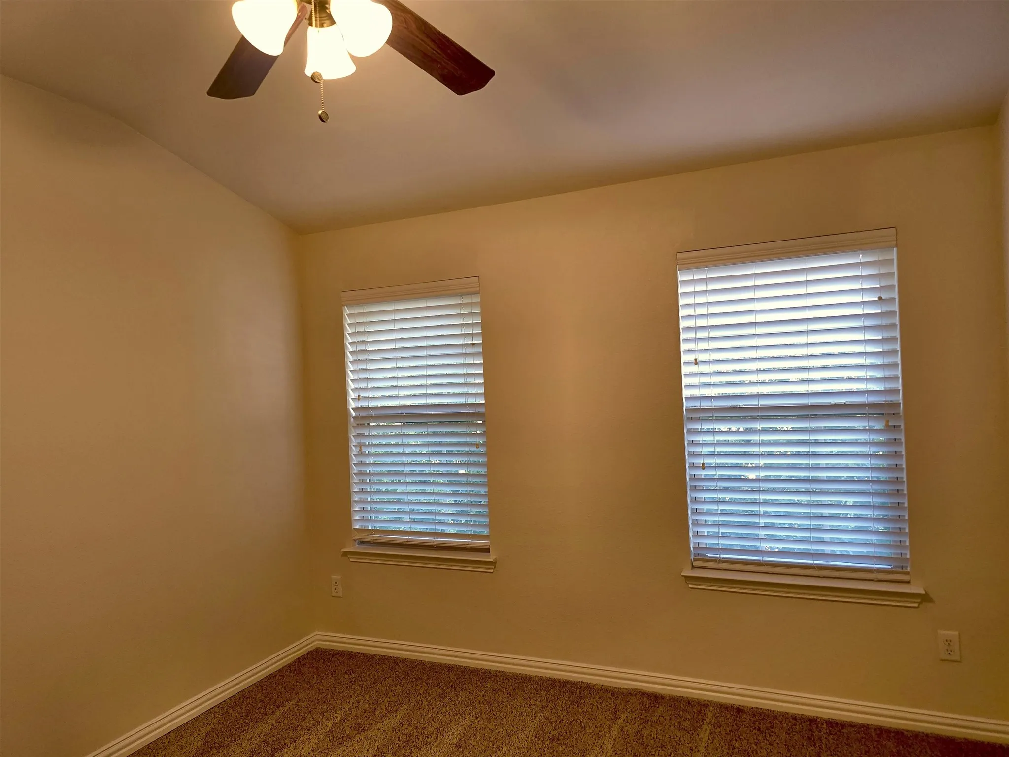 Ceiling fan with integrated lighting, two windows with blinds, light-colored walls, and carpet flooring