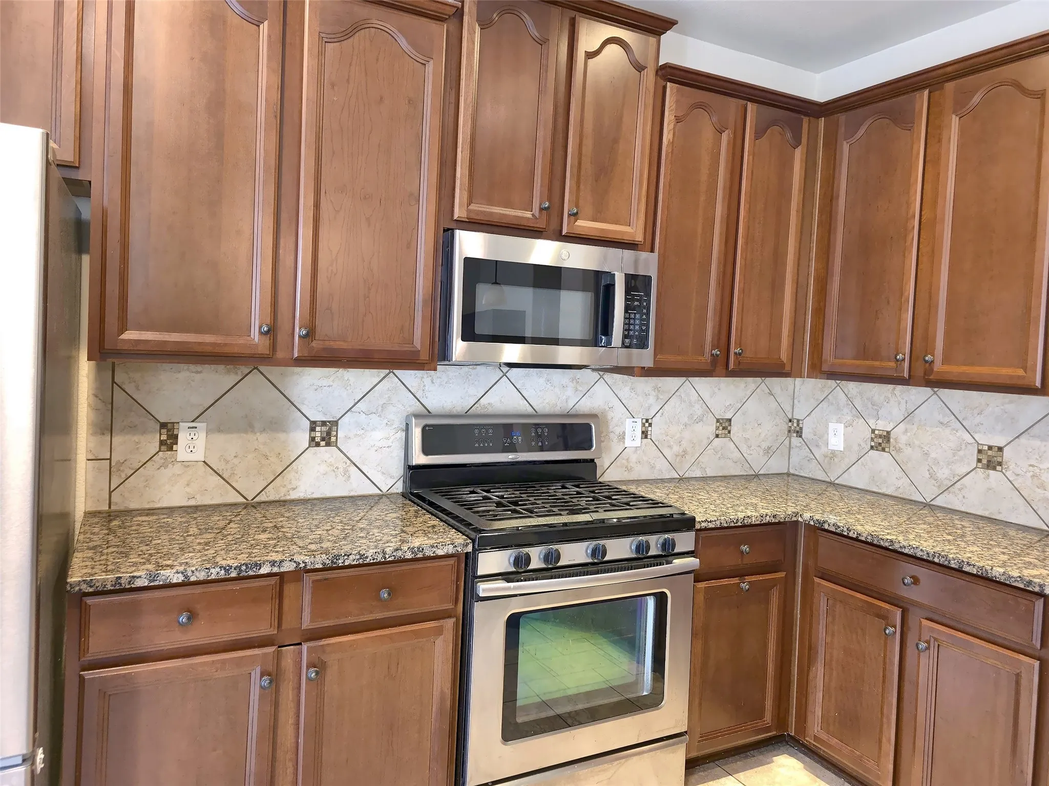 Dark wood cabinetry, stainless steel appliances, tile backsplash with decorative accents, and speckled countertops