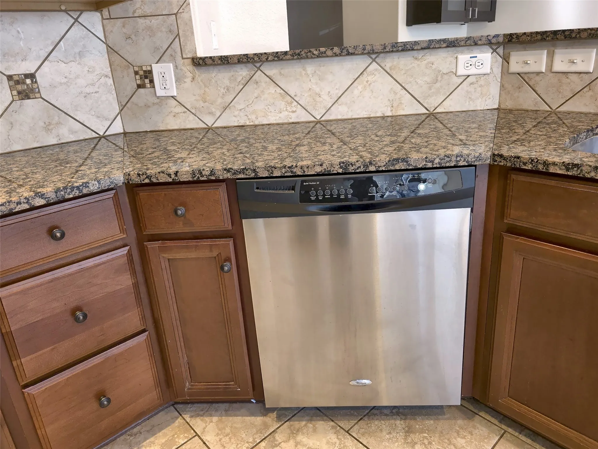 Stainless steel dishwasher, granite-style countertops, wood-finish cabinetry, tile backsplash with decorative inserts, and tile flooring