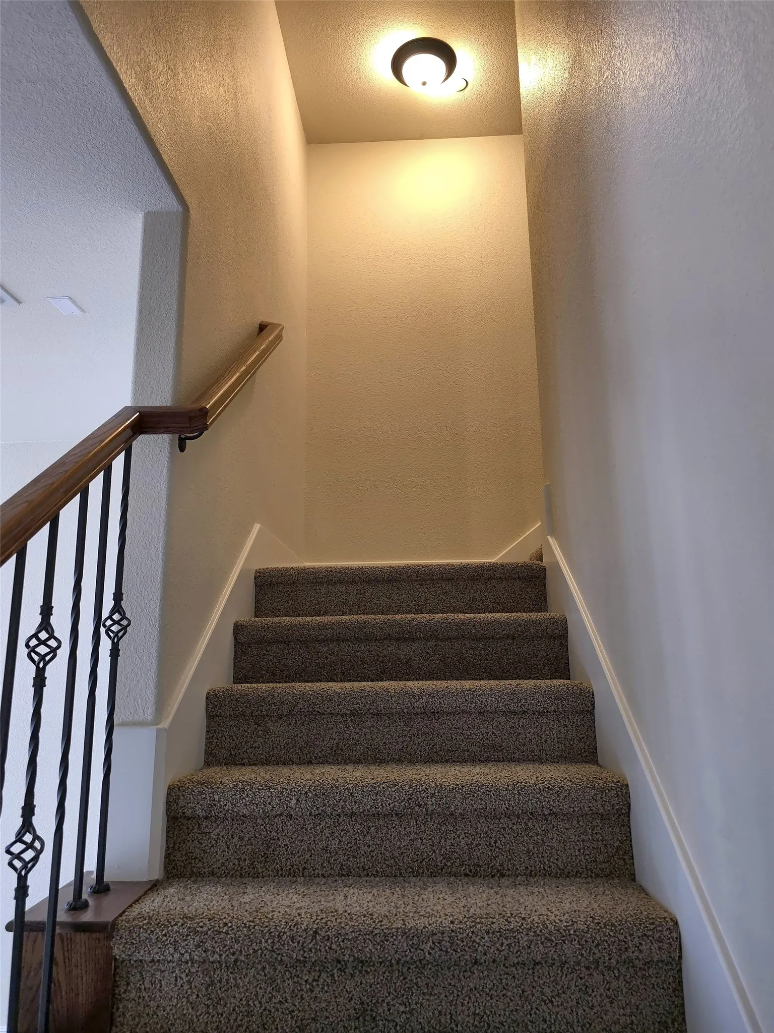 Carpeted staircase featuring wrought iron balusters with decorative twists, a wood-finish handrail, and a flush-mount ceiling light