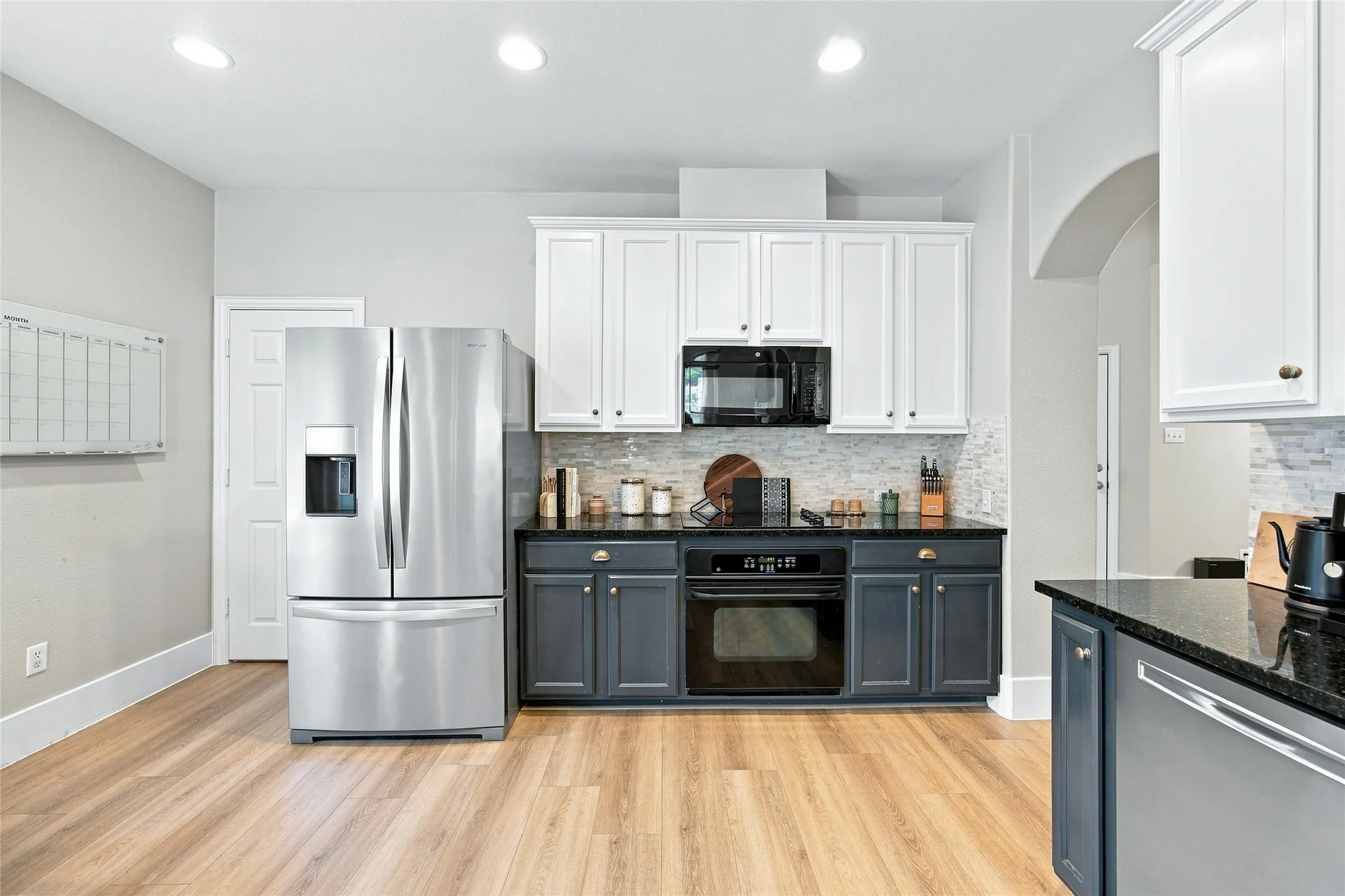Kitchen featuring black countertops, wood-finish flooring, white upper cabinetry, and dark lower cabinetry. The  walk in pantry is located just next to the refridgerator space perfectly tucked away.