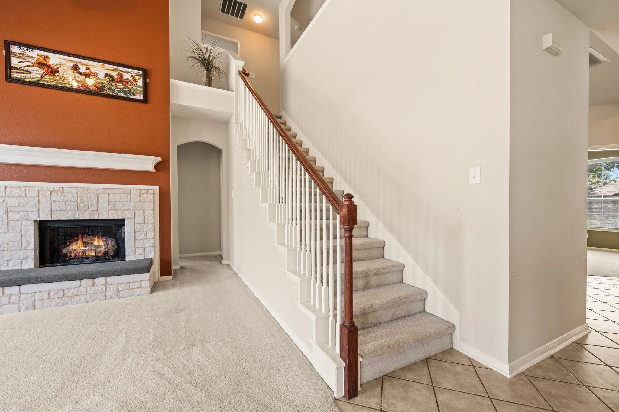 Stone-clad fireplace with mantel, carpeted flooring, staircase with wood handrail and white balusters, arched entryway, and tiled flooring