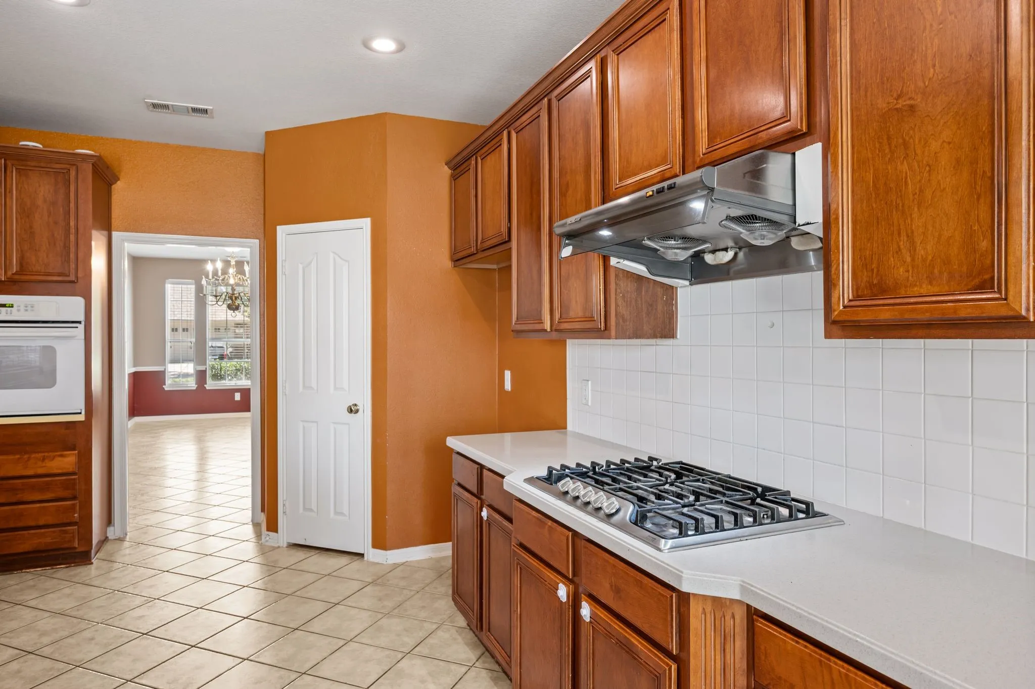 Wood-finish cabinetry, built-in oven, integrated gas cooktop, tile flooring, and a white tile backsplash