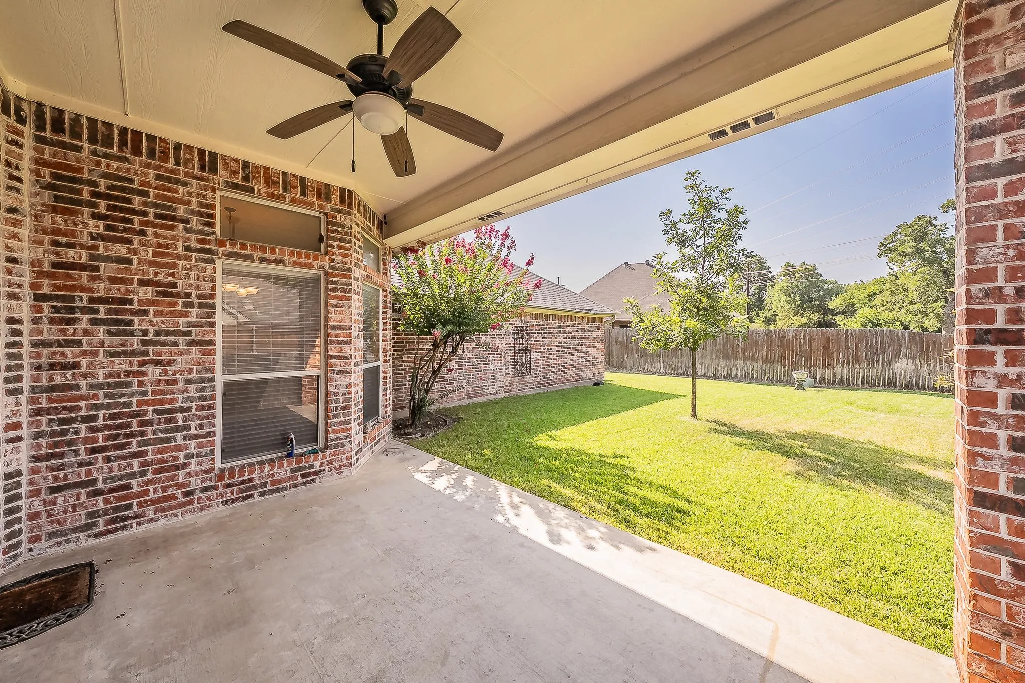 Covered patio featuring a ceiling fan, brick exterior, concrete flooring, and an expansive lawn with a wooden privacy fence