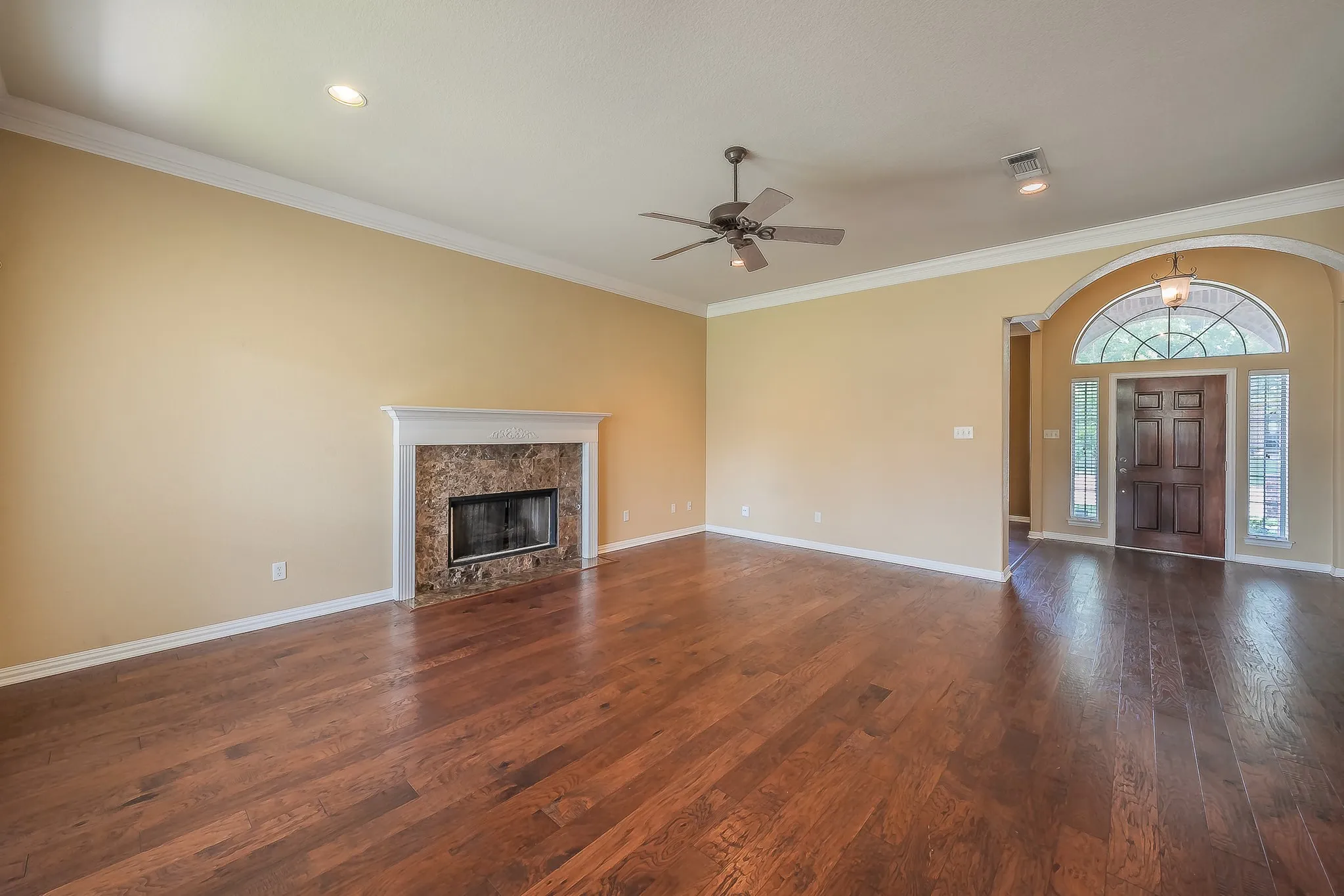 Wood-finish flooring, fireplace with stone surround, crown molding, arched entryway, and a dark wood front door with sidelights