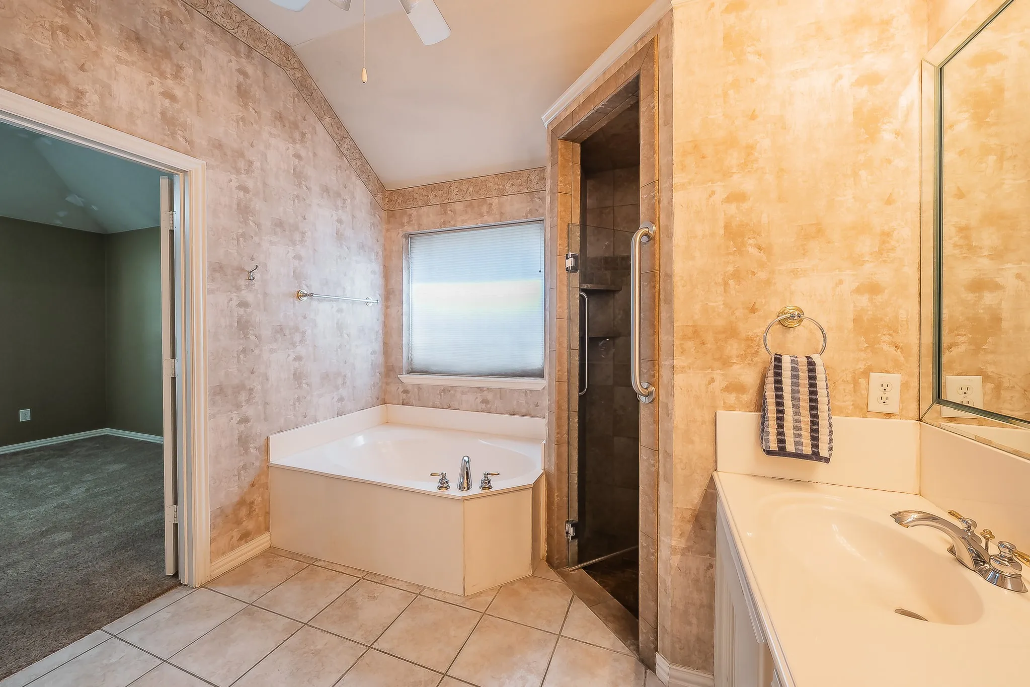 Corner soaking tub, separate glass-enclosed shower, single vanity with integrated sink, ceramic tile flooring, and vaulted ceiling with ceiling fan