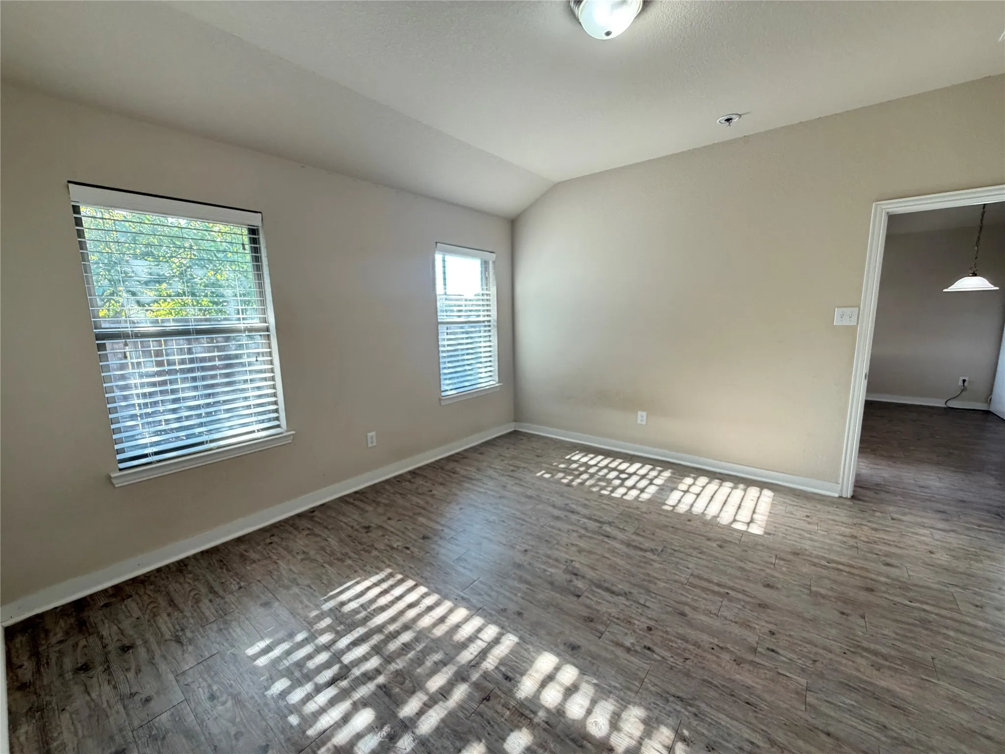 Vaulted ceiling, two windows with blinds, wood-finish flooring, and a flush mount ceiling light