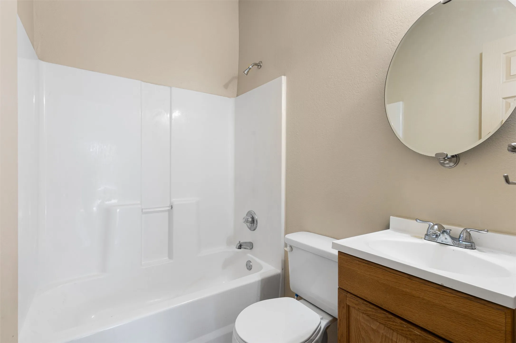 White bathtub and shower surround with chrome fixtures, wood-finish vanity with white countertop and sink, and a circular mirror