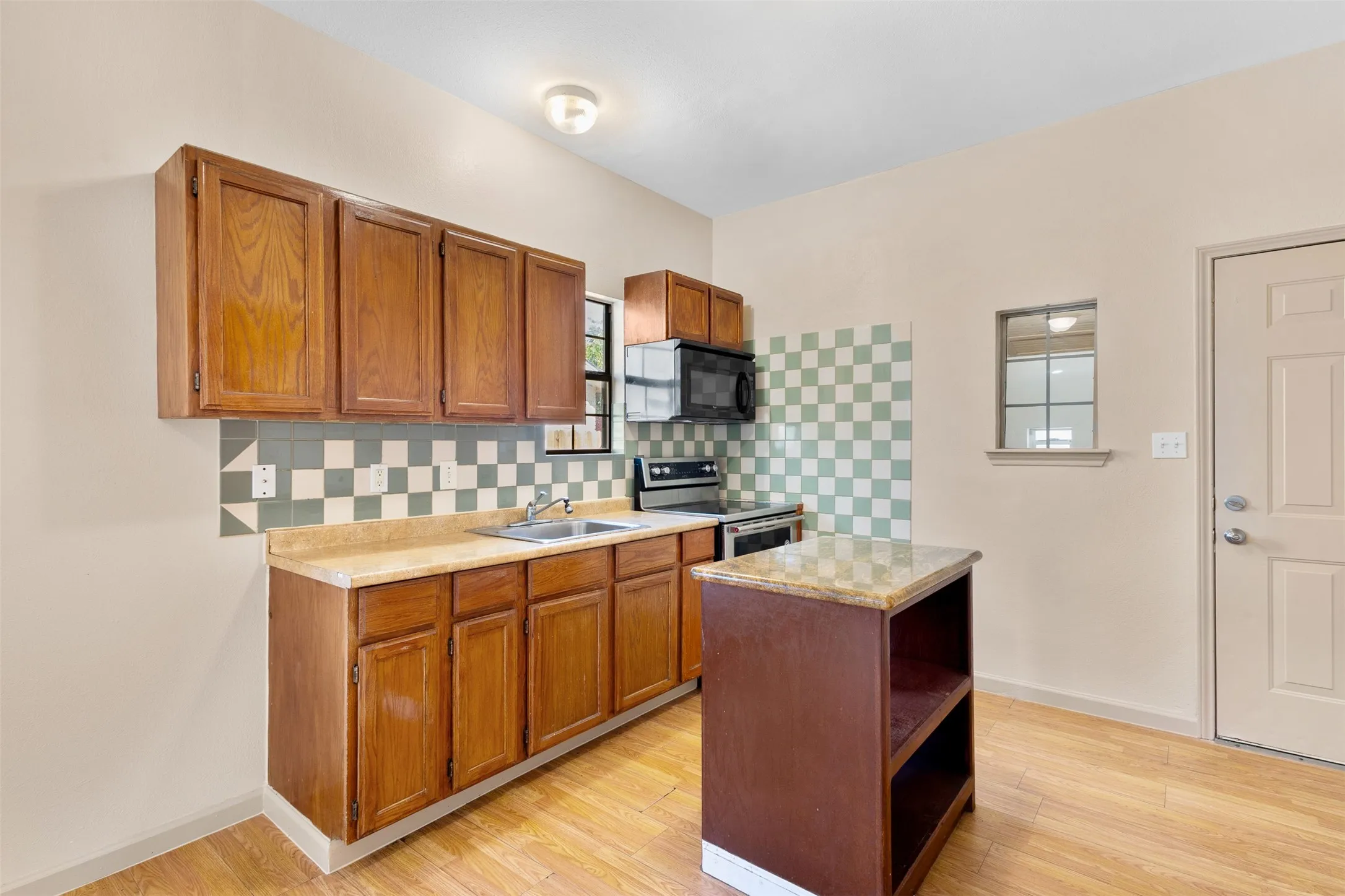 Wood-finish flooring, wood cabinetry, built-in microwave, checkerboard tile backsplash, and a kitchen island with a stone-finish countertop