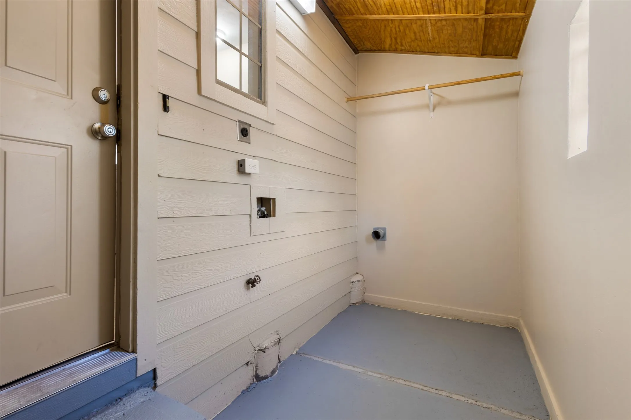 Utility space featuring a wood-panel wall, a single window, a built-in hanging rod, and a concrete floor