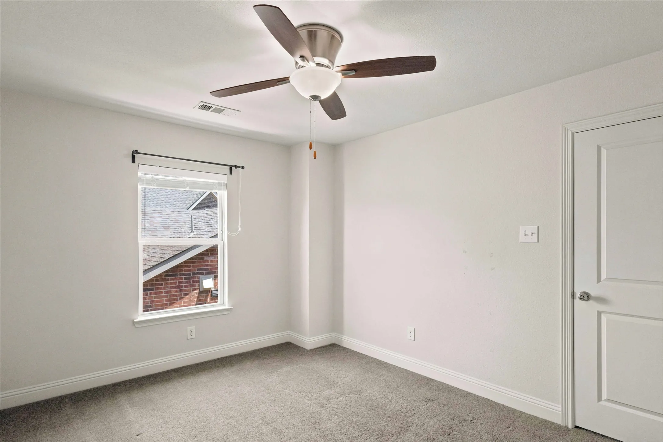 Ceiling fan with wood-finish blades and integrated lighting, light gray wall paint, window with white trim, white paneled door, and gray carpeting