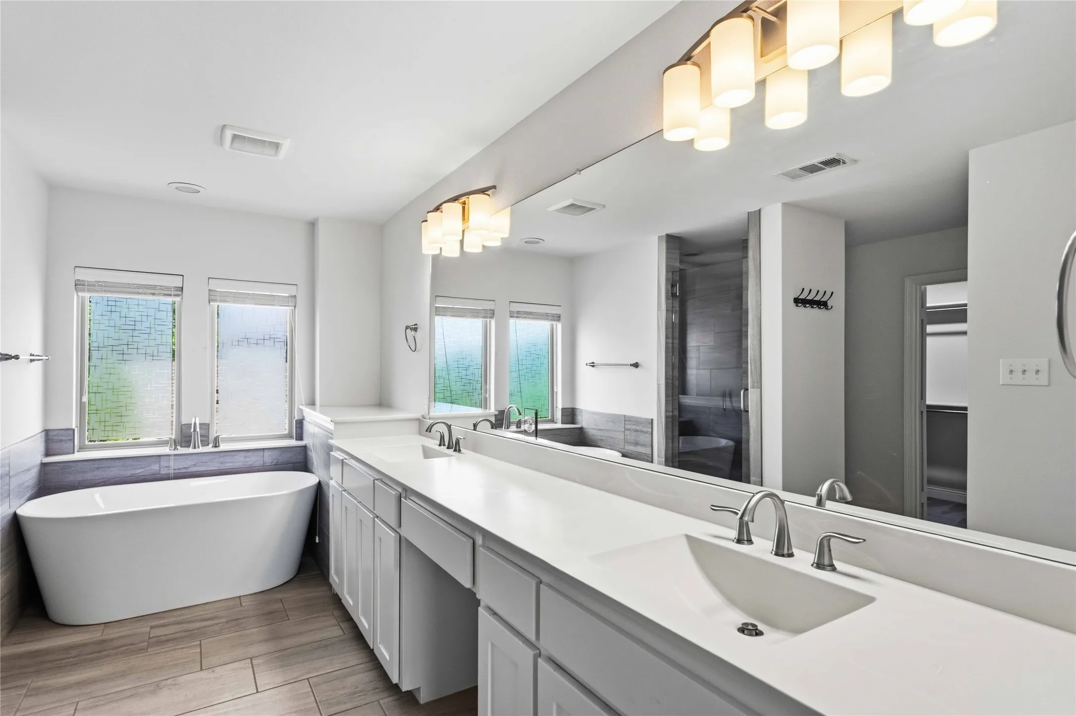 Dual vanity with integrated sinks and brushed nickel fixtures, expansive mirror, wood-finish flooring, freestanding soaking tub, and a separate shower enclosure