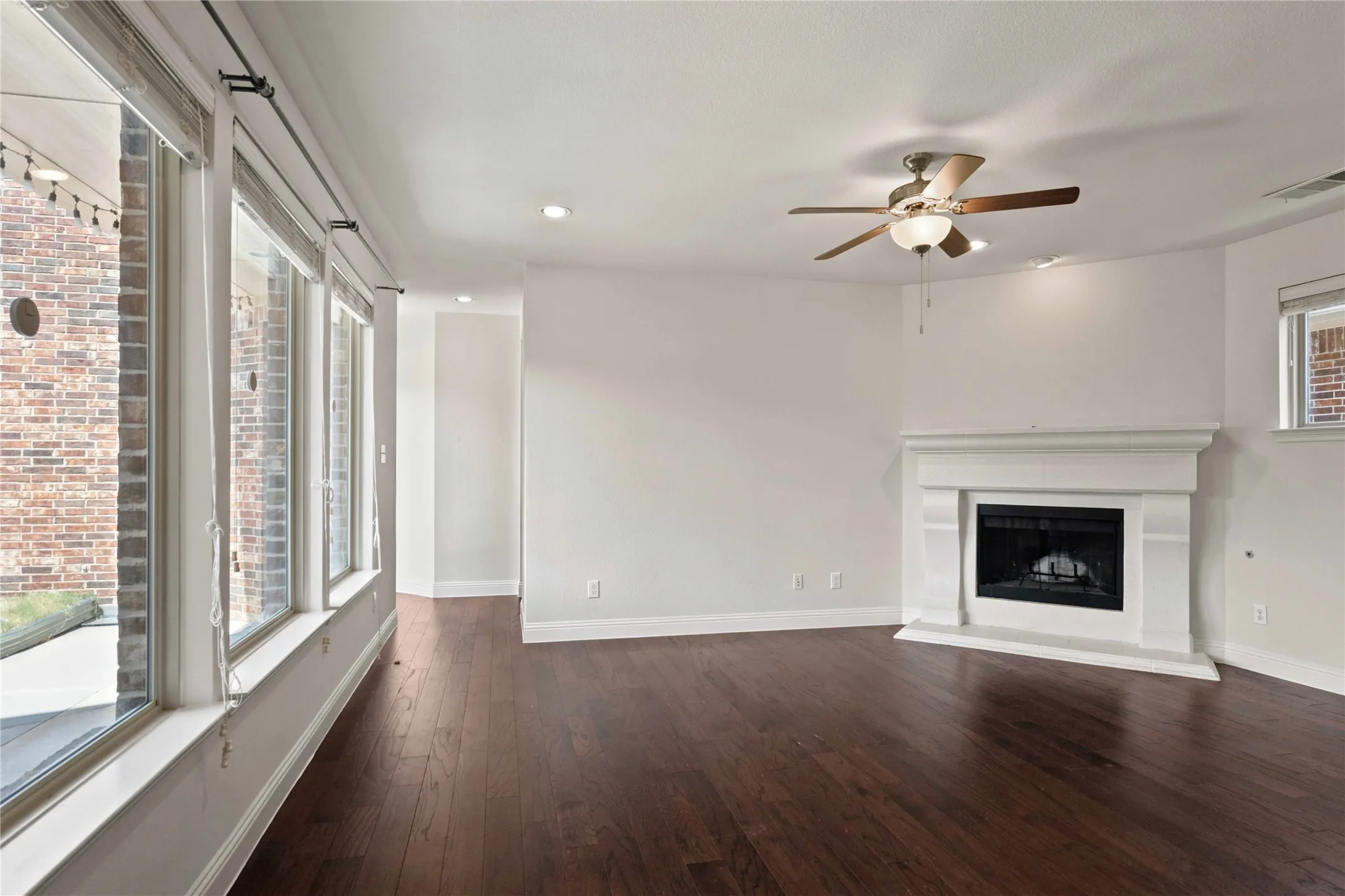 Dark wood-finish flooring, white fireplace with hearth, ceiling fan with integrated lighting, recessed lighting, and a wall of windows