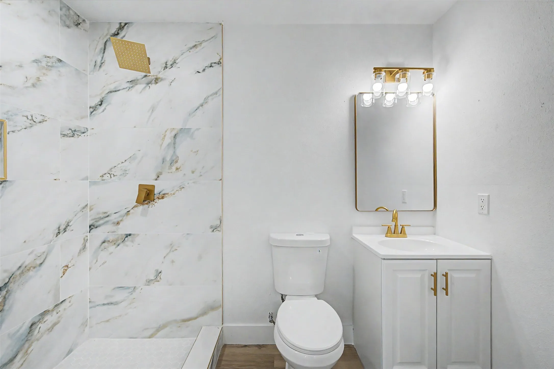 Walk-in shower with large format marble-look tile, gold-tone fixtures, white vanity with gold-tone hardware, and a framed mirror with an overhead light fixture