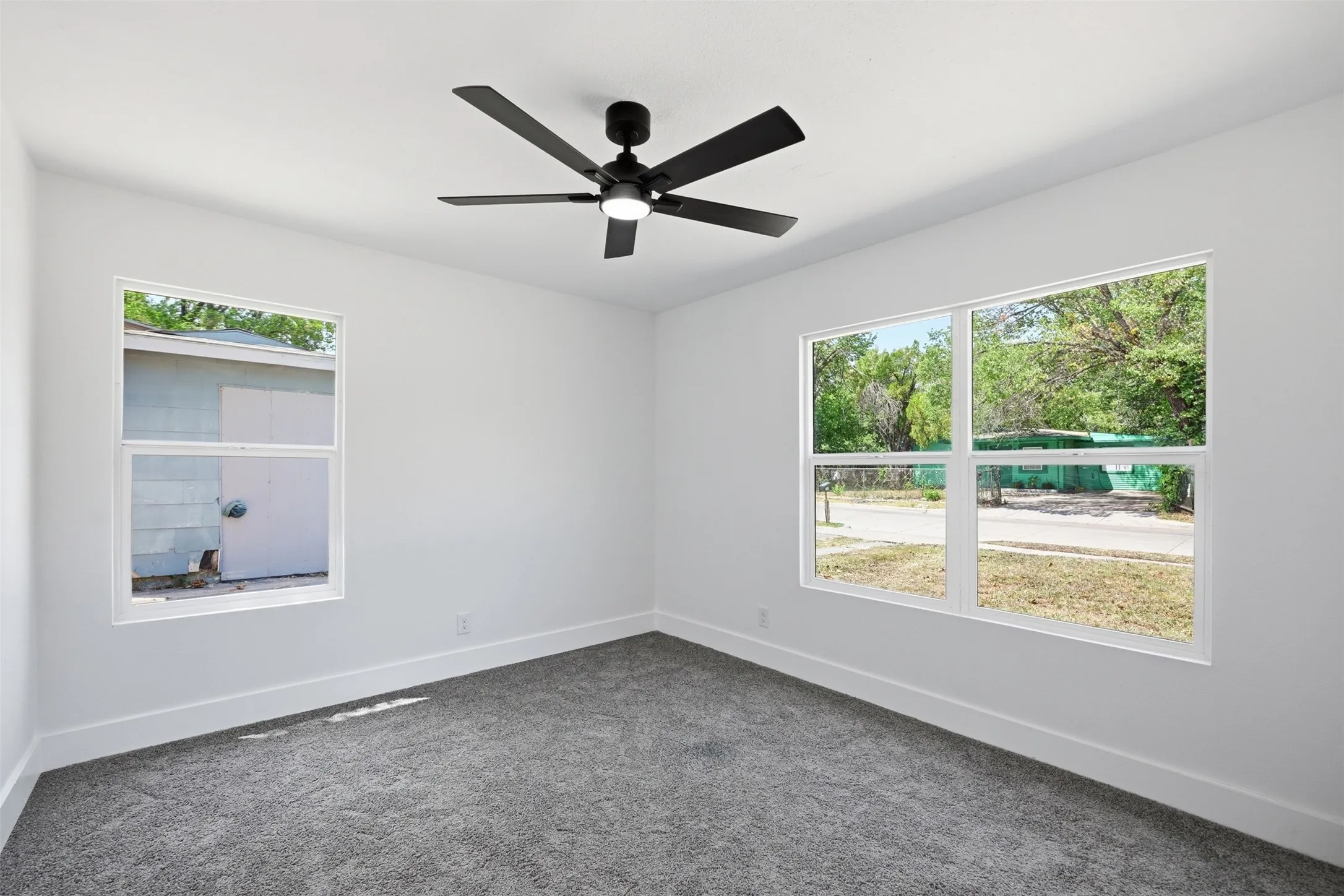 Crisp white walls, plush gray carpeting, modern ceiling fan with integrated lighting, and multiple windows providing ample illumination