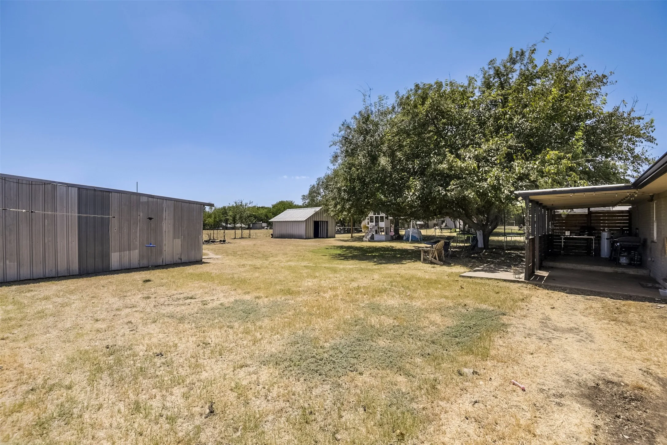 Expansive outdoor area with a large mature tree, a covered patio structure, and a secondary building with a pitched roof