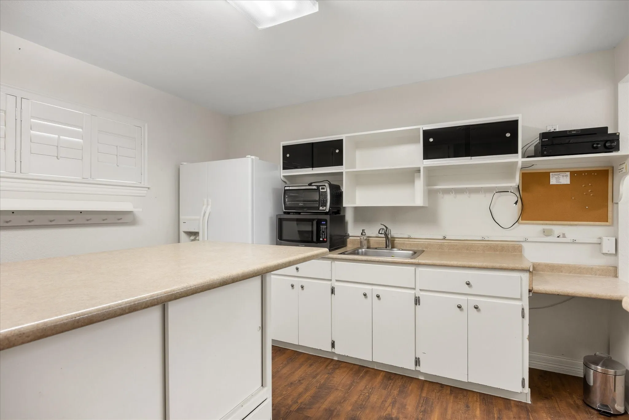White cabinetry with silver hardware, laminate countertops, stainless steel sink, wood-finish flooring, and white plantation shutters