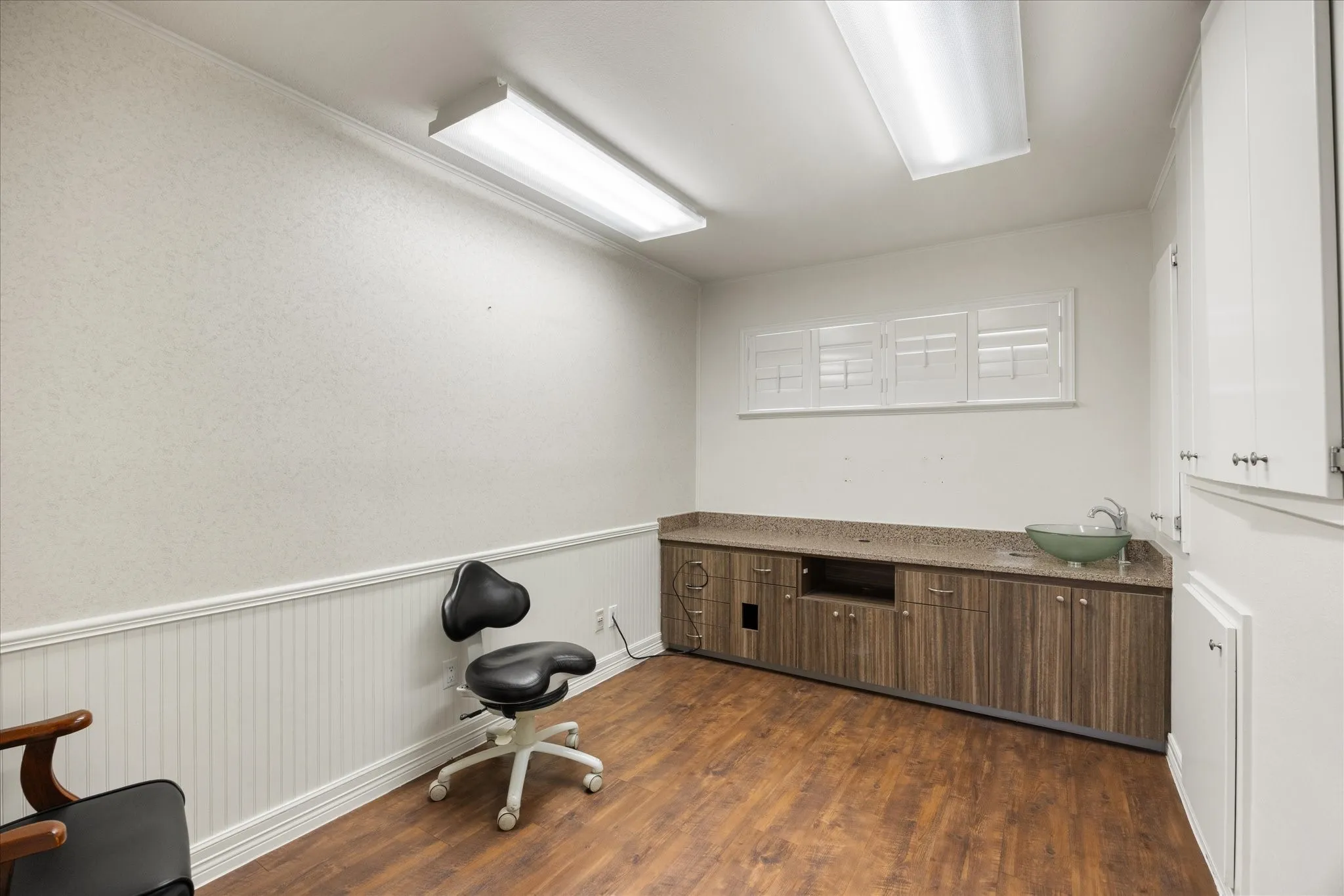 Wood-finish flooring, white wainscoting, built-in cabinetry with a stone-finish countertop, and a vessel sink