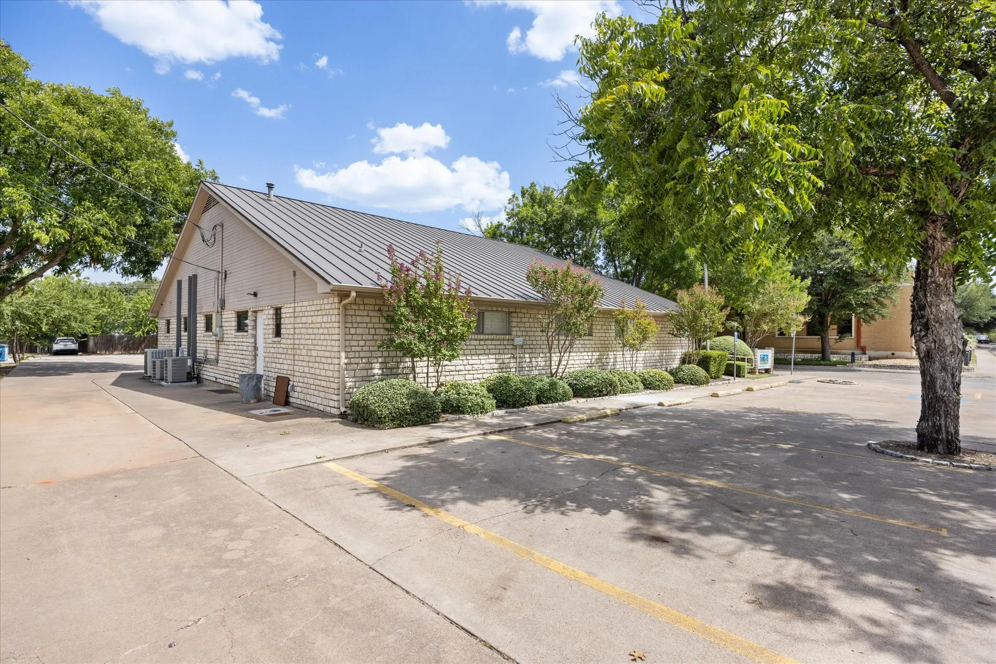 Exterior featuring a brick facade, pitched metal roof, paved parking area, and manicured landscaping