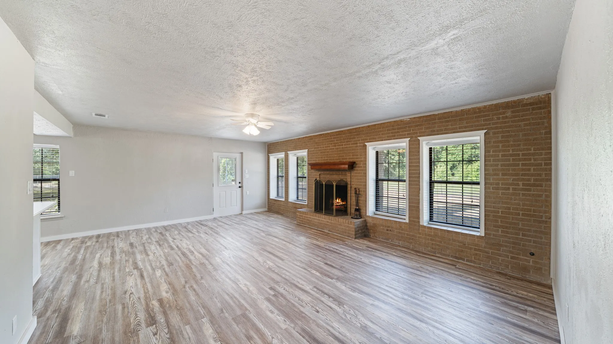Spacious room featuring wood-finish flooring, a brick accent wall with multiple windows, and a built-in fireplace with a wooden mantel