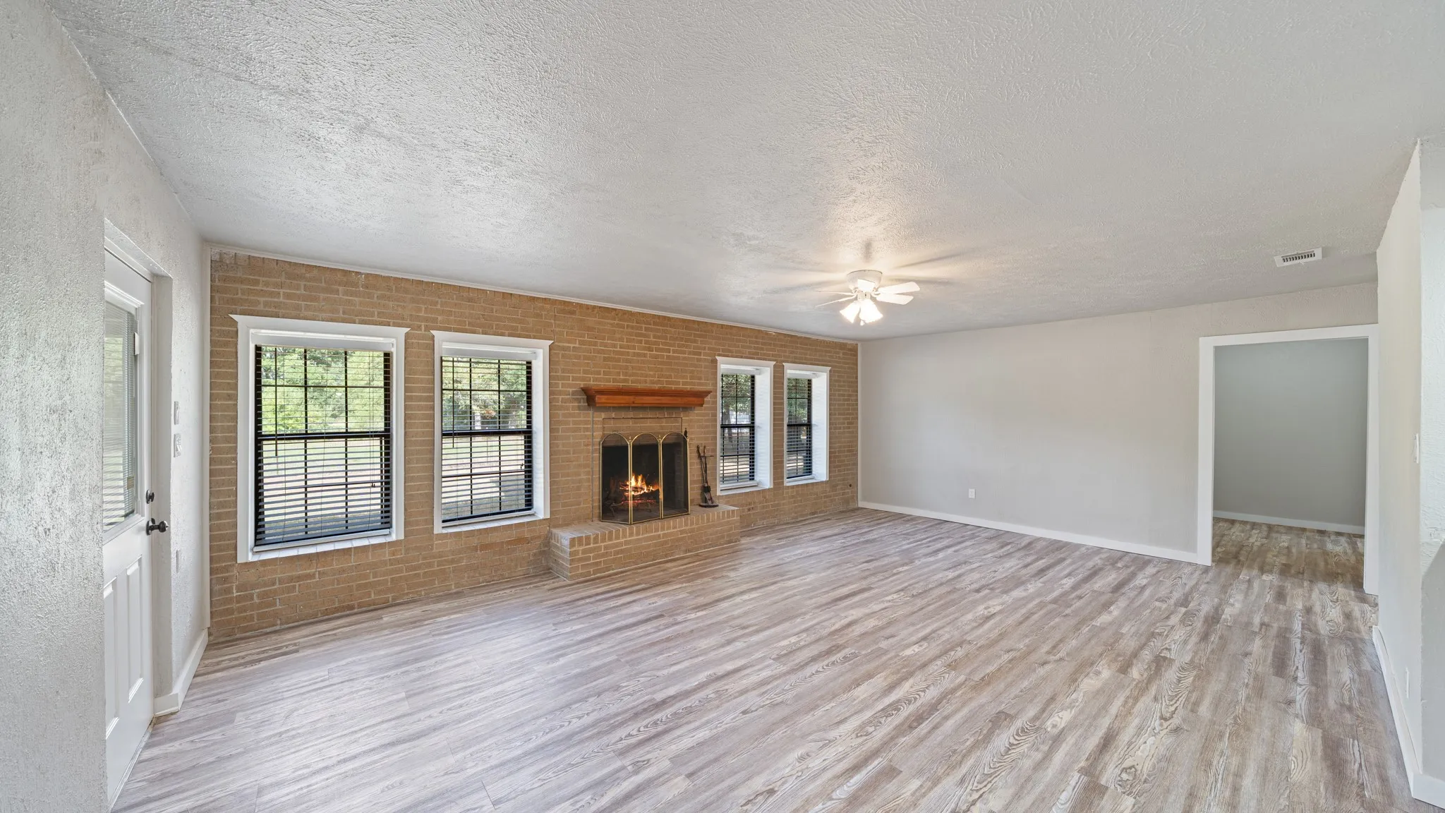 Spacious living area featuring wood-finish flooring, a brick fireplace with a wood mantel, two windows, a ceiling fan, and painted walls