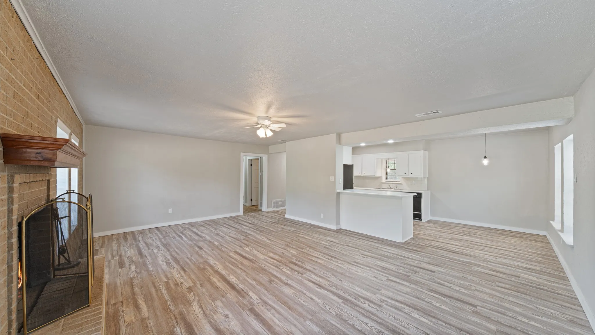 Light wood-finish flooring throughout, brick fireplace with wood mantel, ceiling fan, recessed lighting, and kitchen with white cabinetry and dark appliances